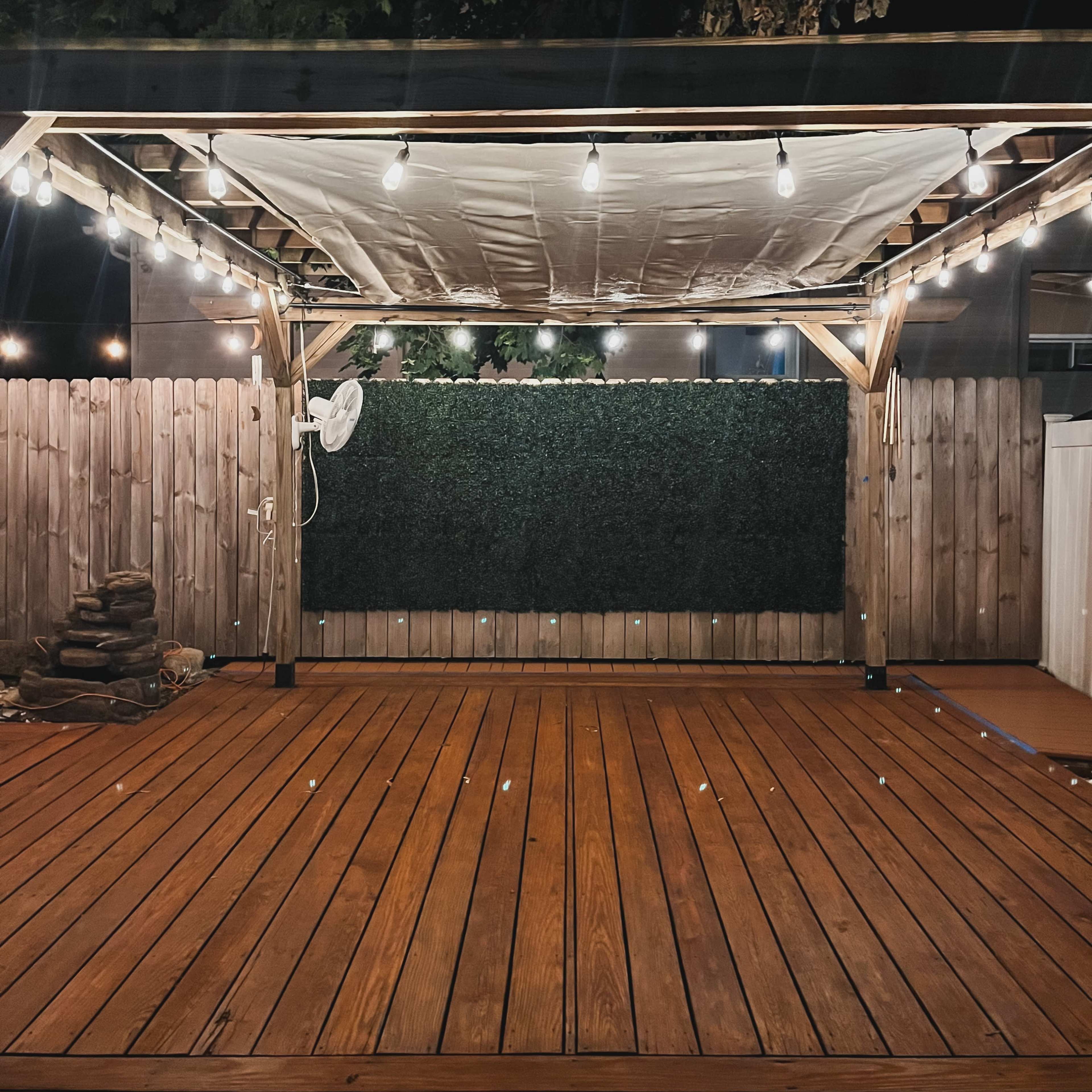 A wooden deck features a pergola with string lights and a white canopy, surrounded by a fenced area with a green backdrop.