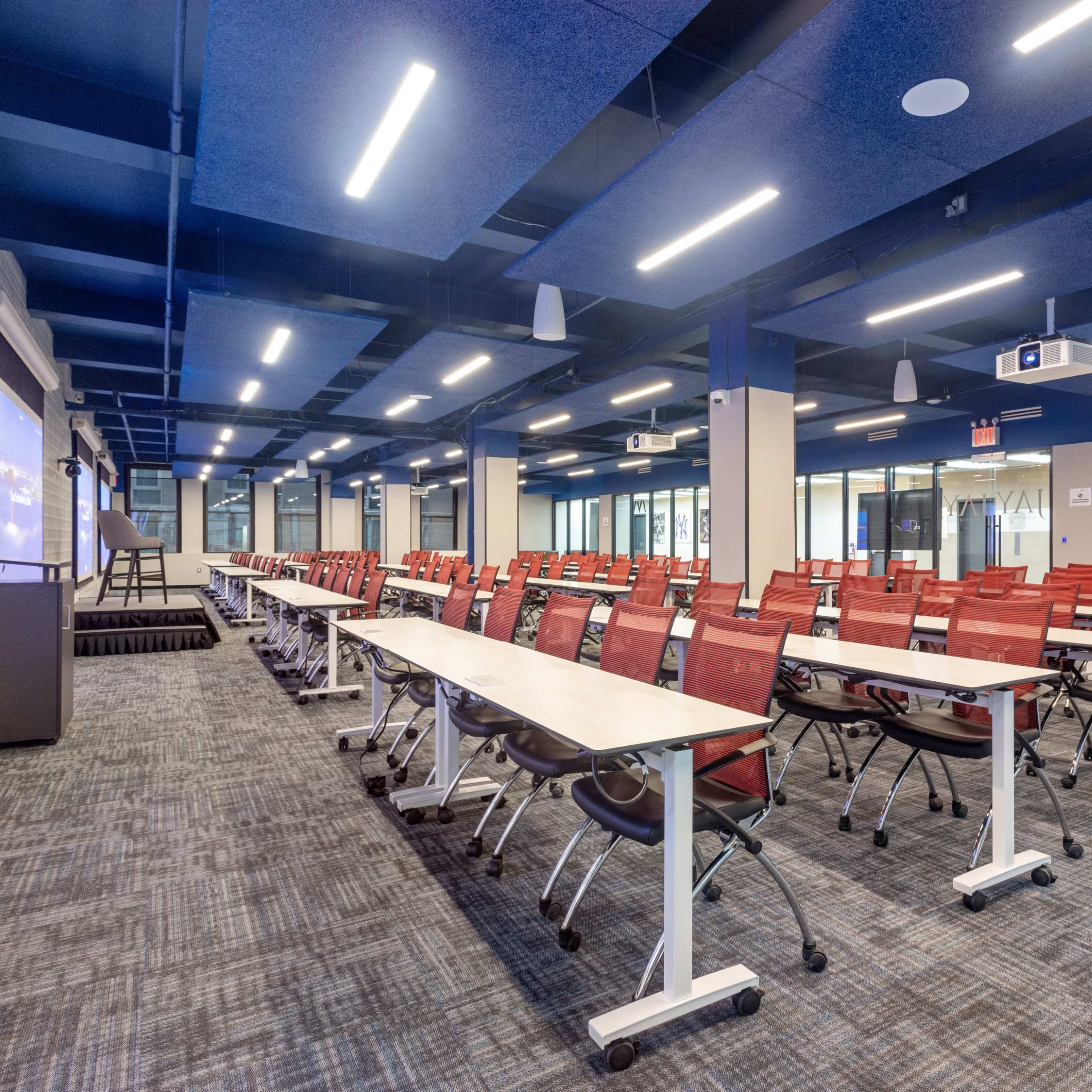 A modern conference room with rows of red chairs and tables arranged for a meeting, featuring blue ceiling panels and overhead lighting.