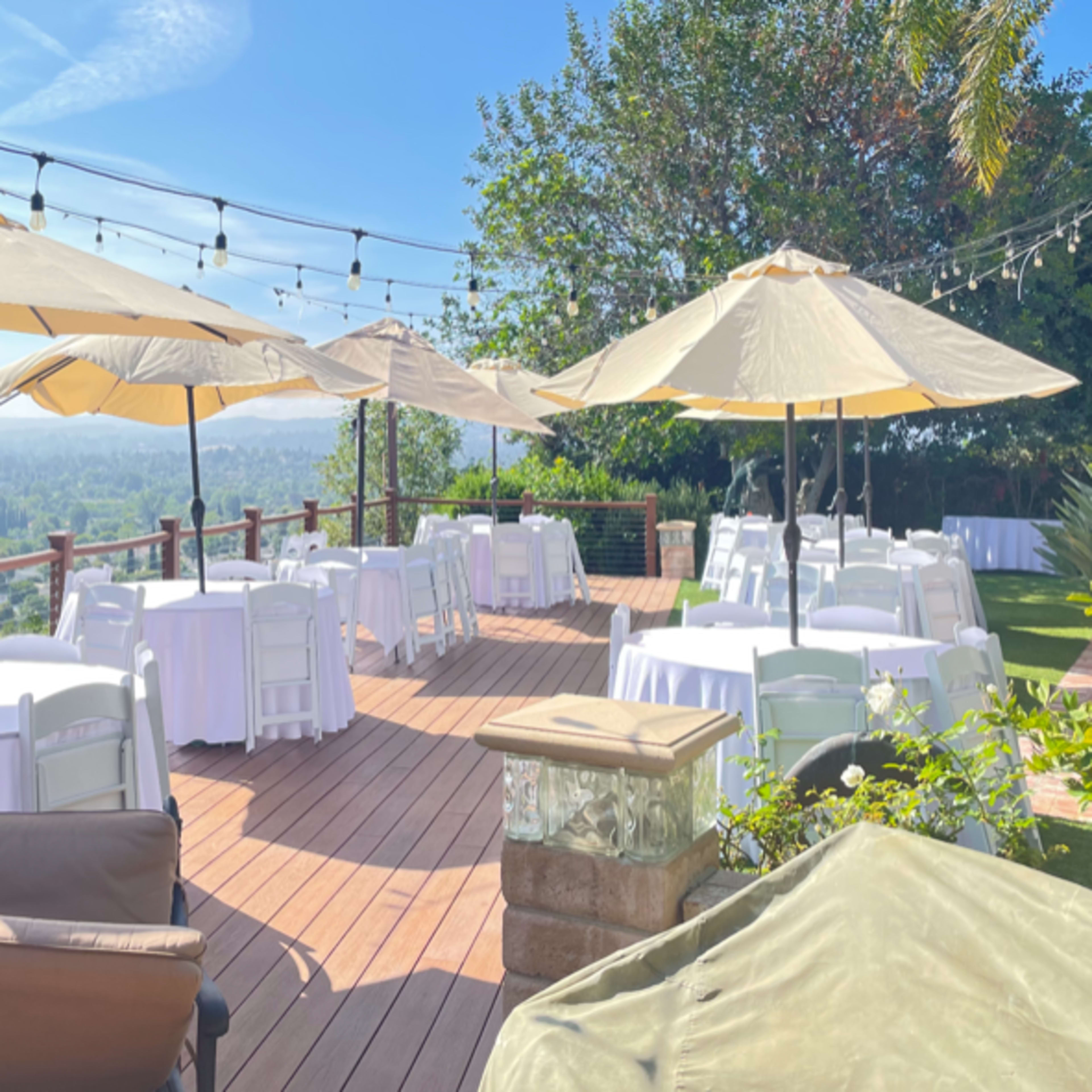 A deck area arranged with white tables and umbrellas, overlooking a scenic landscape under a clear blue sky.