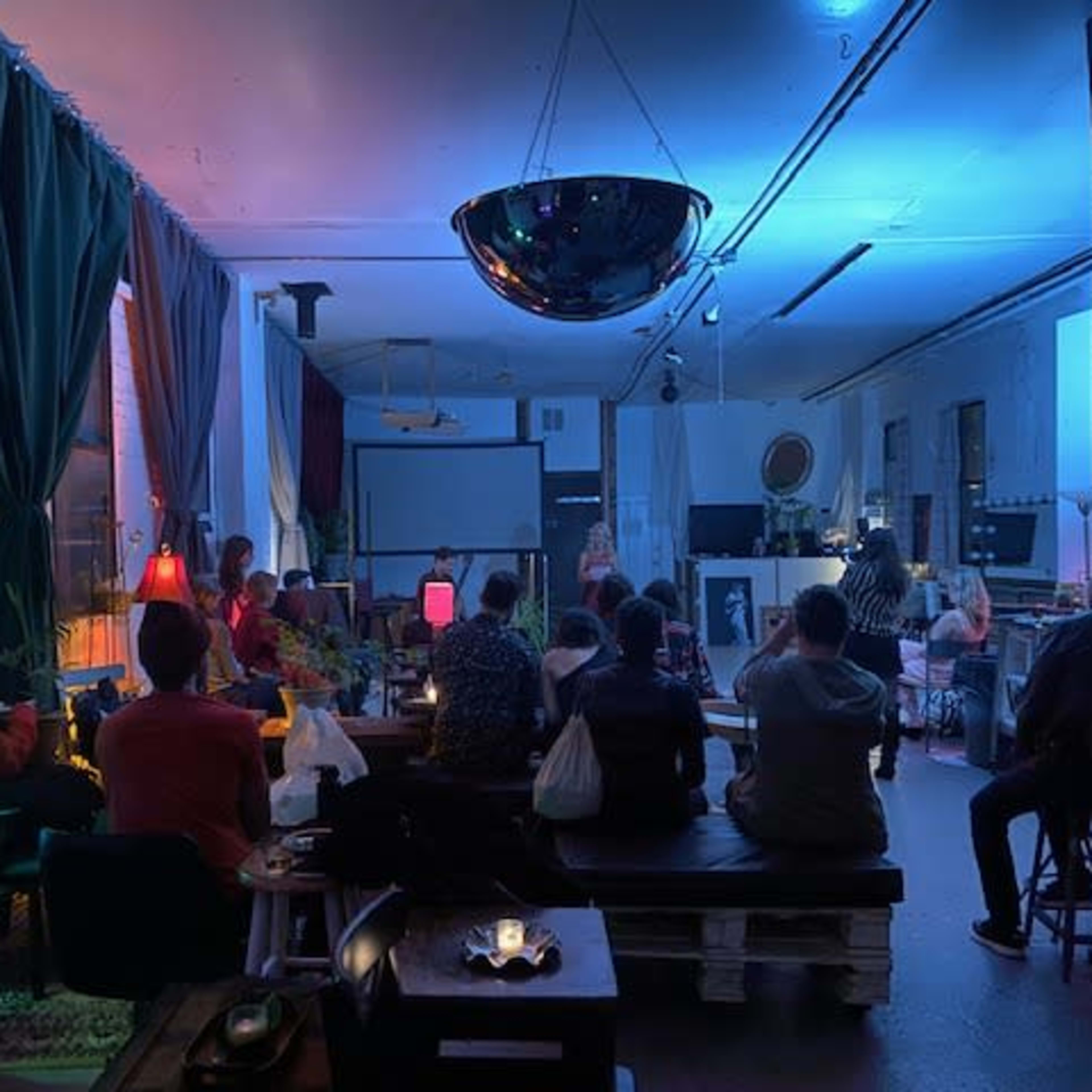 A group of people sit in a dimly lit room with colorful lamps during a gathering or presentation.