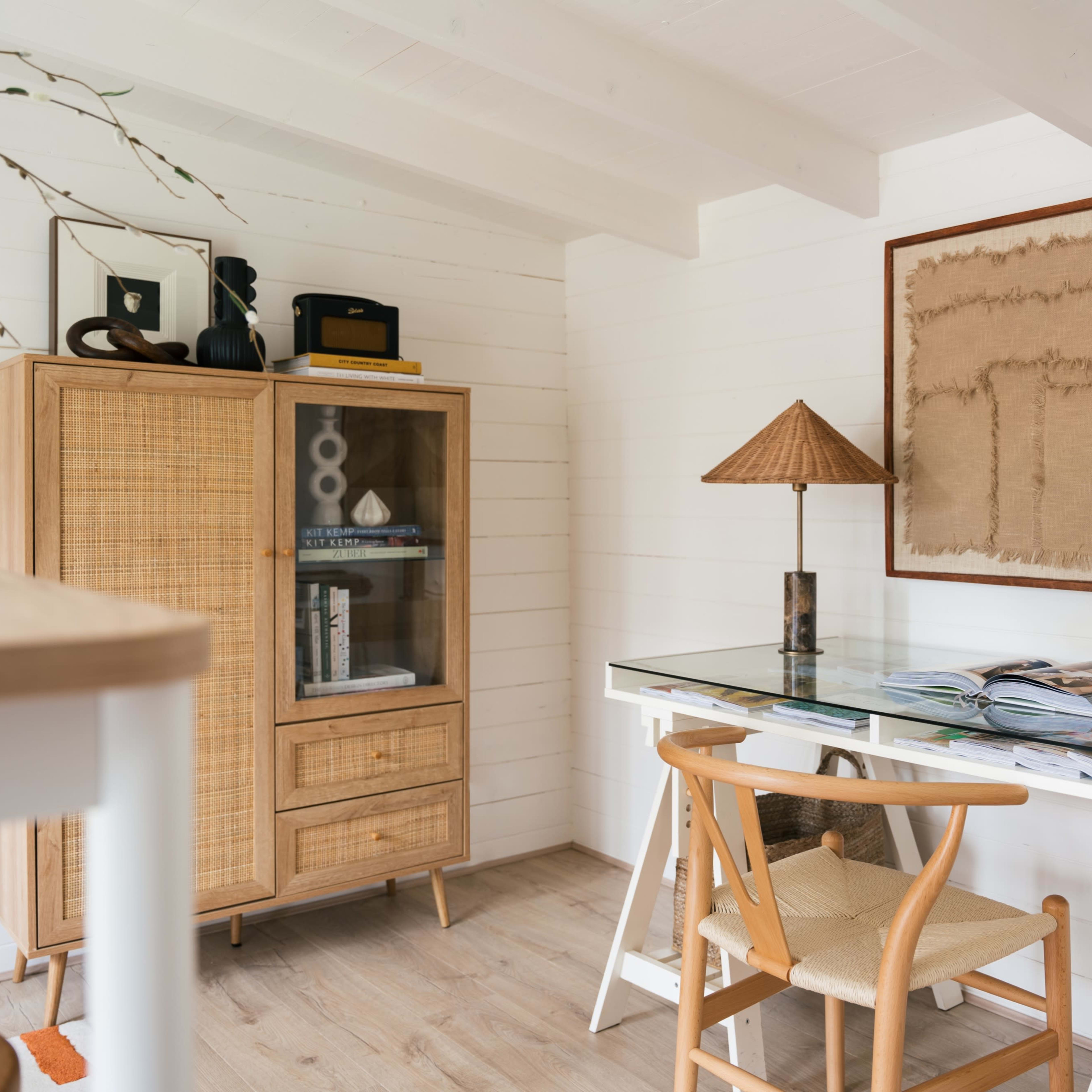 A wooden desk with a glass top, accompanied by a chair, is positioned next to a wicker cabinet in a well-lit room with white walls and wooden beams.