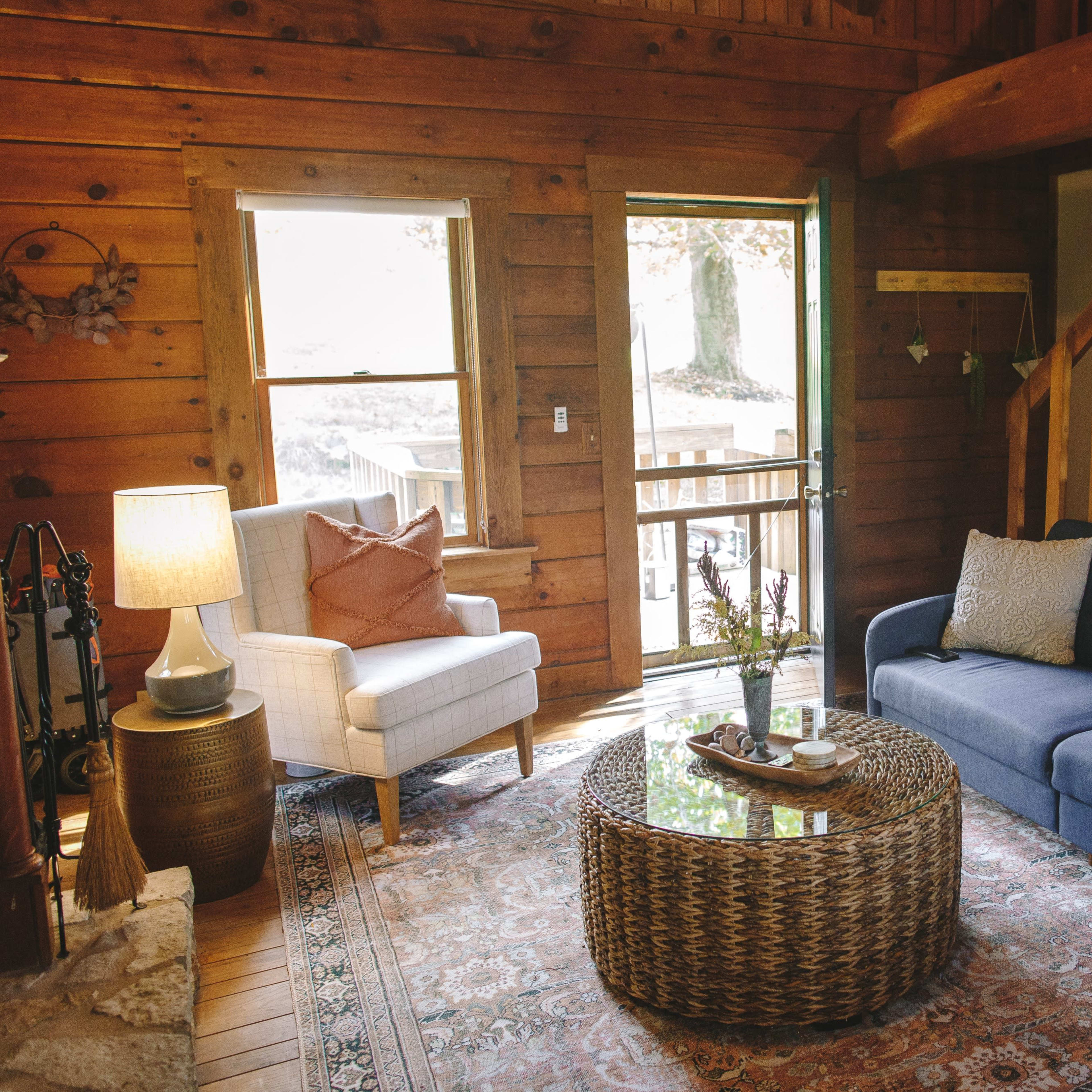 The living room features a blue sofa and a light armchair, a round glass coffee table on a patterned rug, and wooden walls with a staircase in the background.
