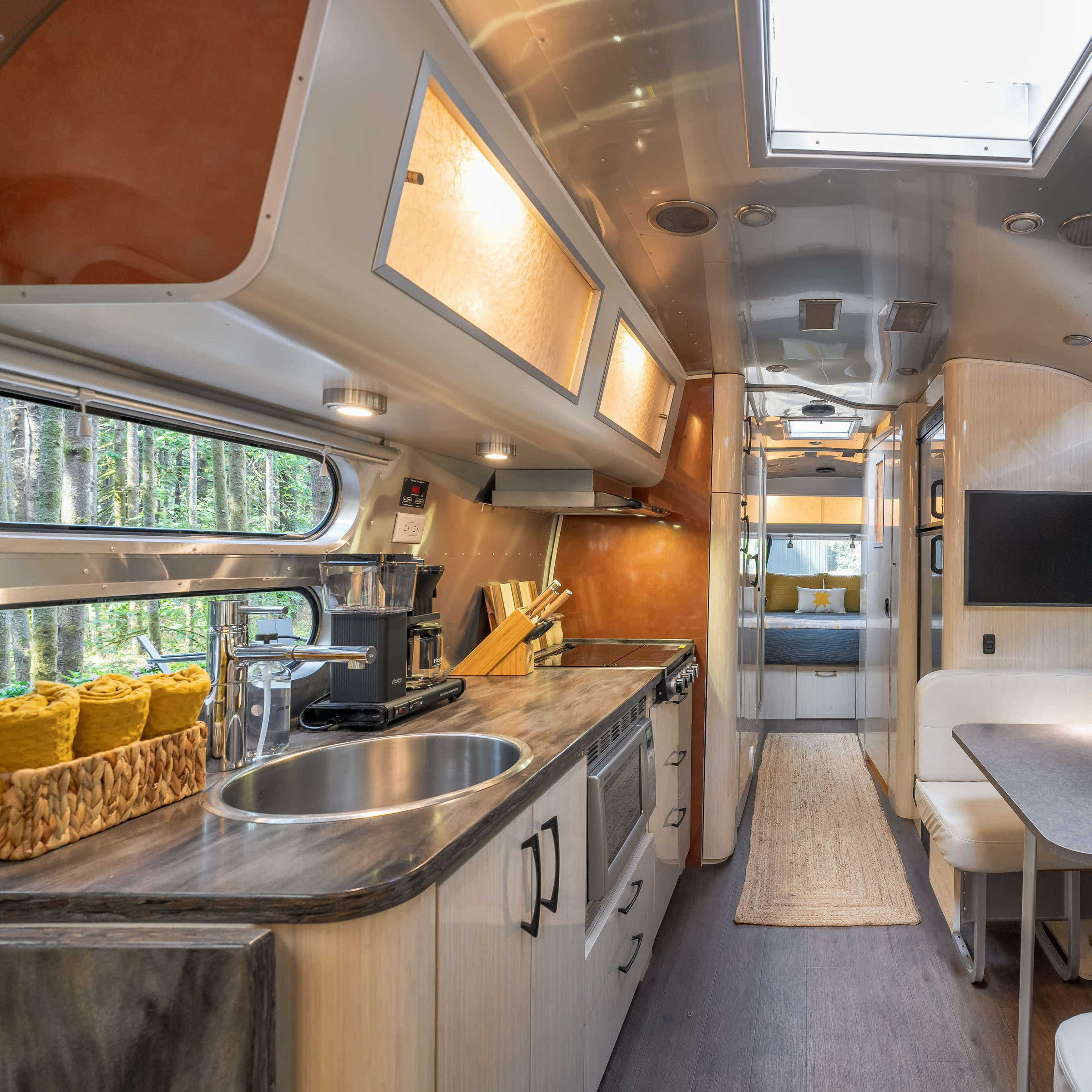 The interior of a modern Airstream trailer features a kitchen area with a circular sink, a dining space with a table, and cozy sleeping quarters visible at the end, all surrounded by large windows.