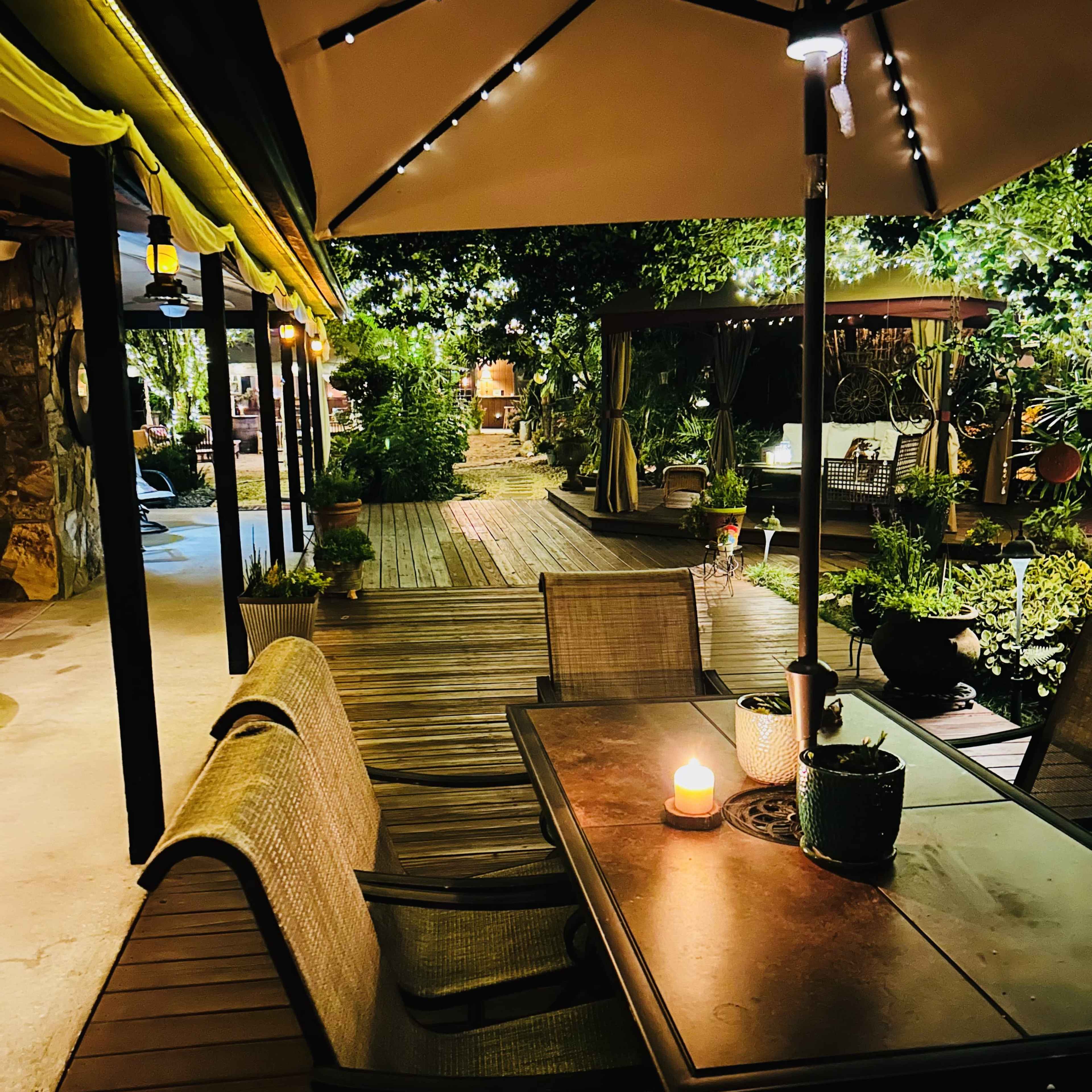 A well-lit outdoor dining area features a table with a candle and chairs beneath an umbrella, surrounded by lush greenery and wooden pathways.
