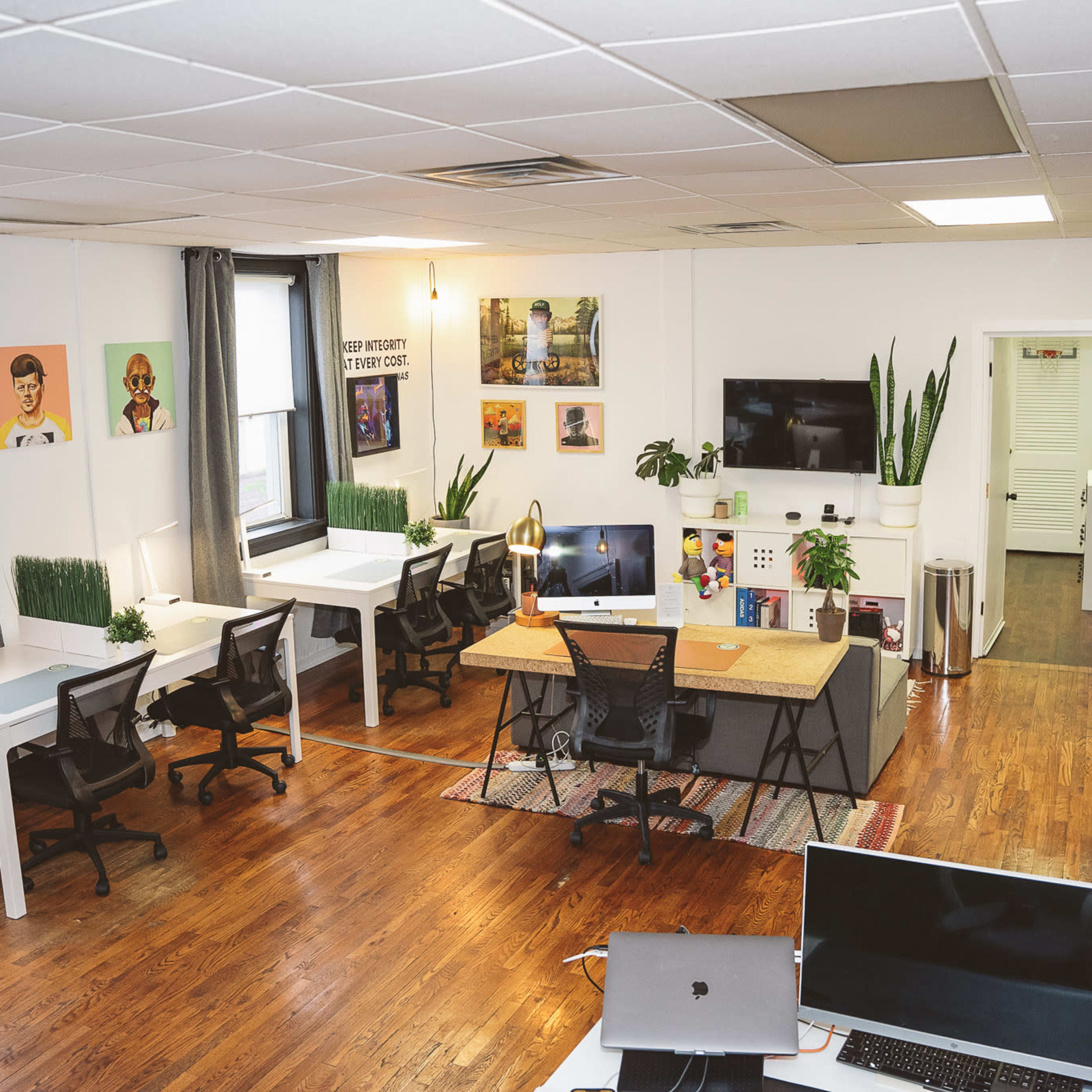 A modern office space with multiple workstations, a central desk, plants, artwork on the walls, and a television mounted on one side.