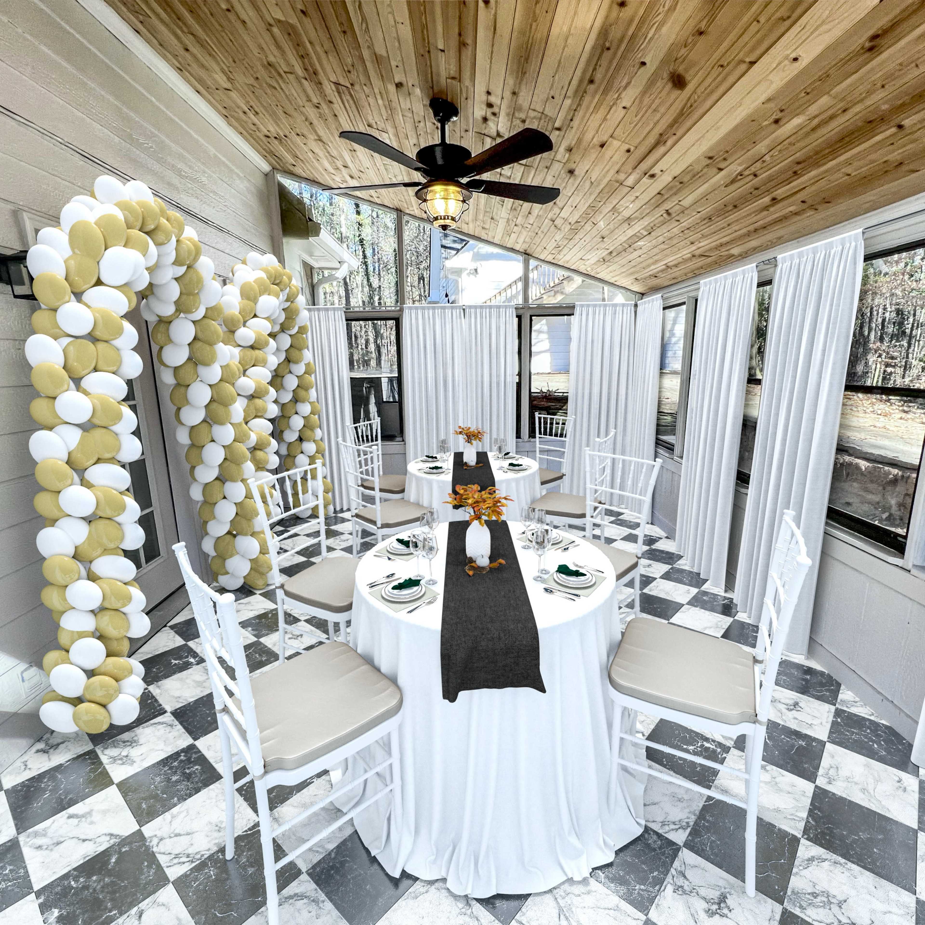 A dining area with a round table set for four, surrounded by white chairs, featuring a decorative balloon arch and large windows against a checkered floor.