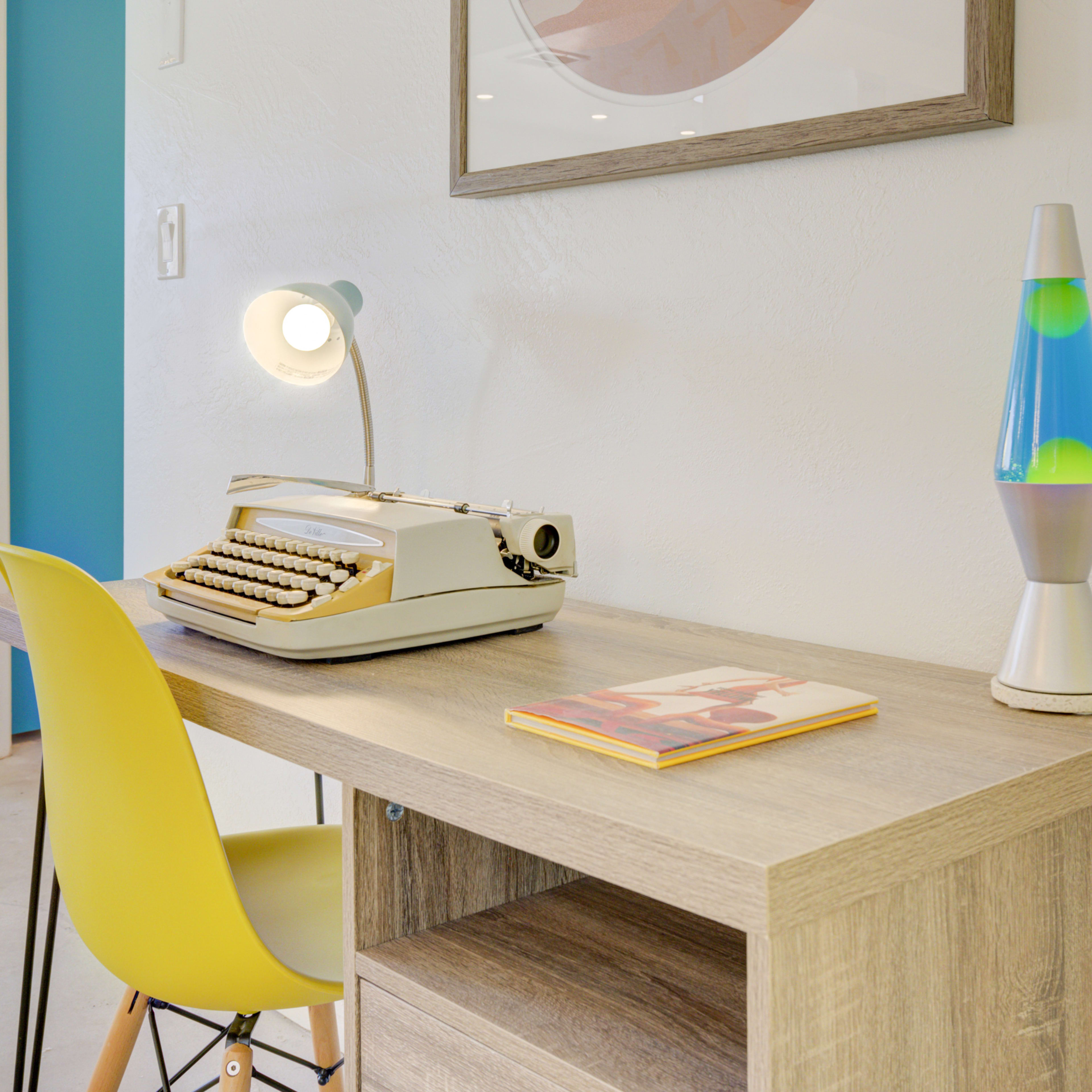 A vintage typewriter sits on a wooden desk next to a colorful lava lamp and a yellow chair.