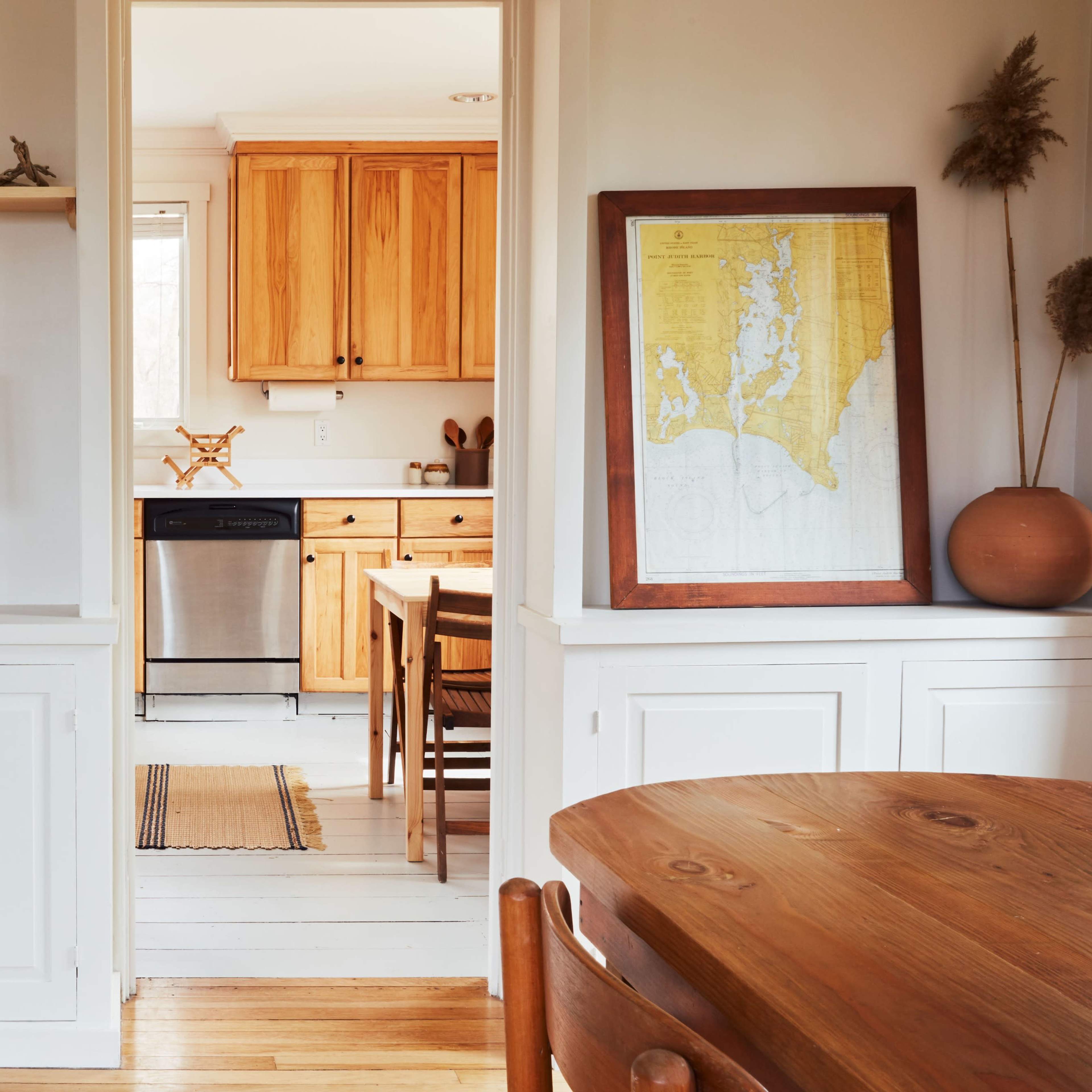 A bright interior space featuring a wooden dining table, a lamp on a shelf, and a view into a kitchen with wooden cabinets and a map on the wall.