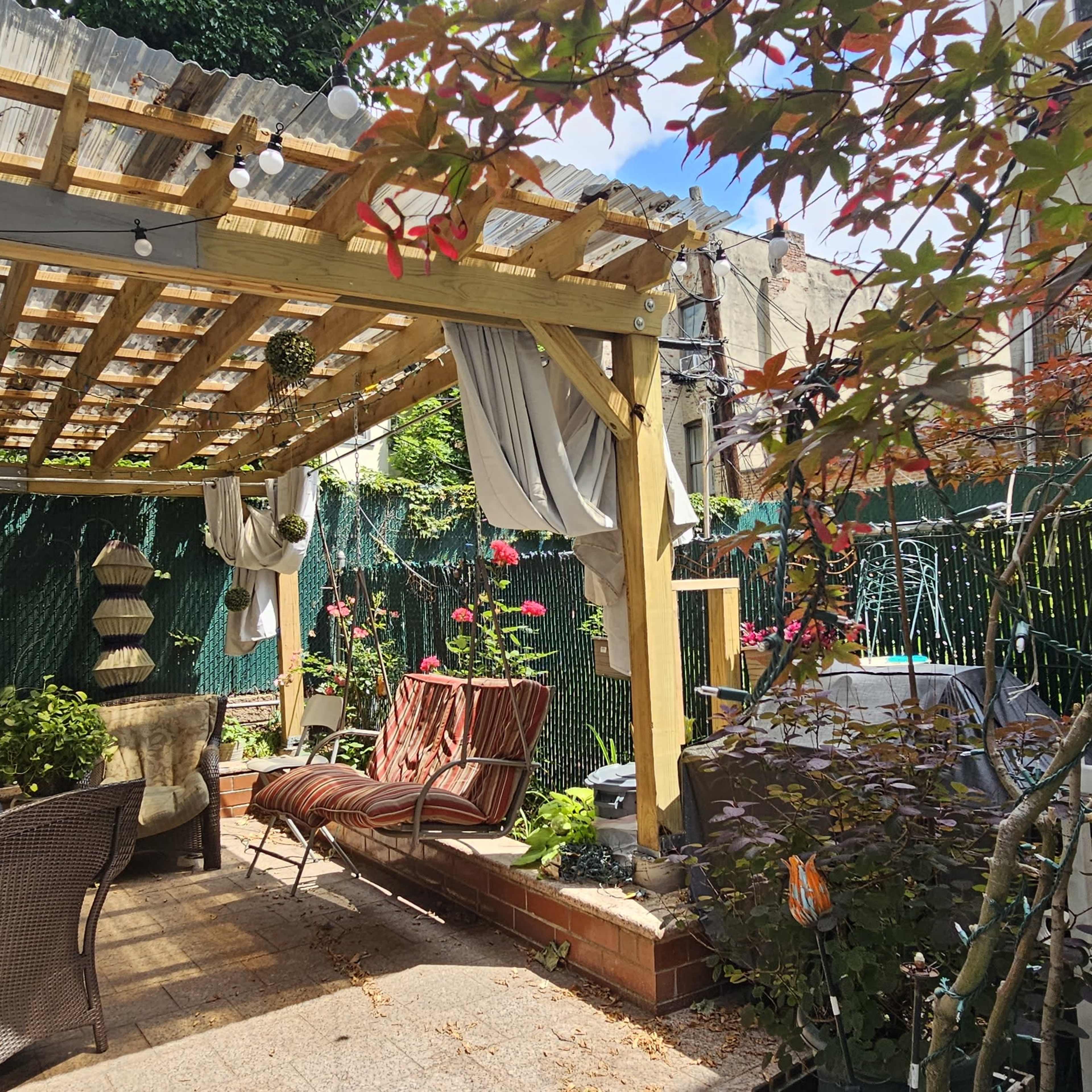 A backyard patio is sheltered by a wooden pergola adorned with hanging plants and string lights, featuring seating areas and surrounding greenery.