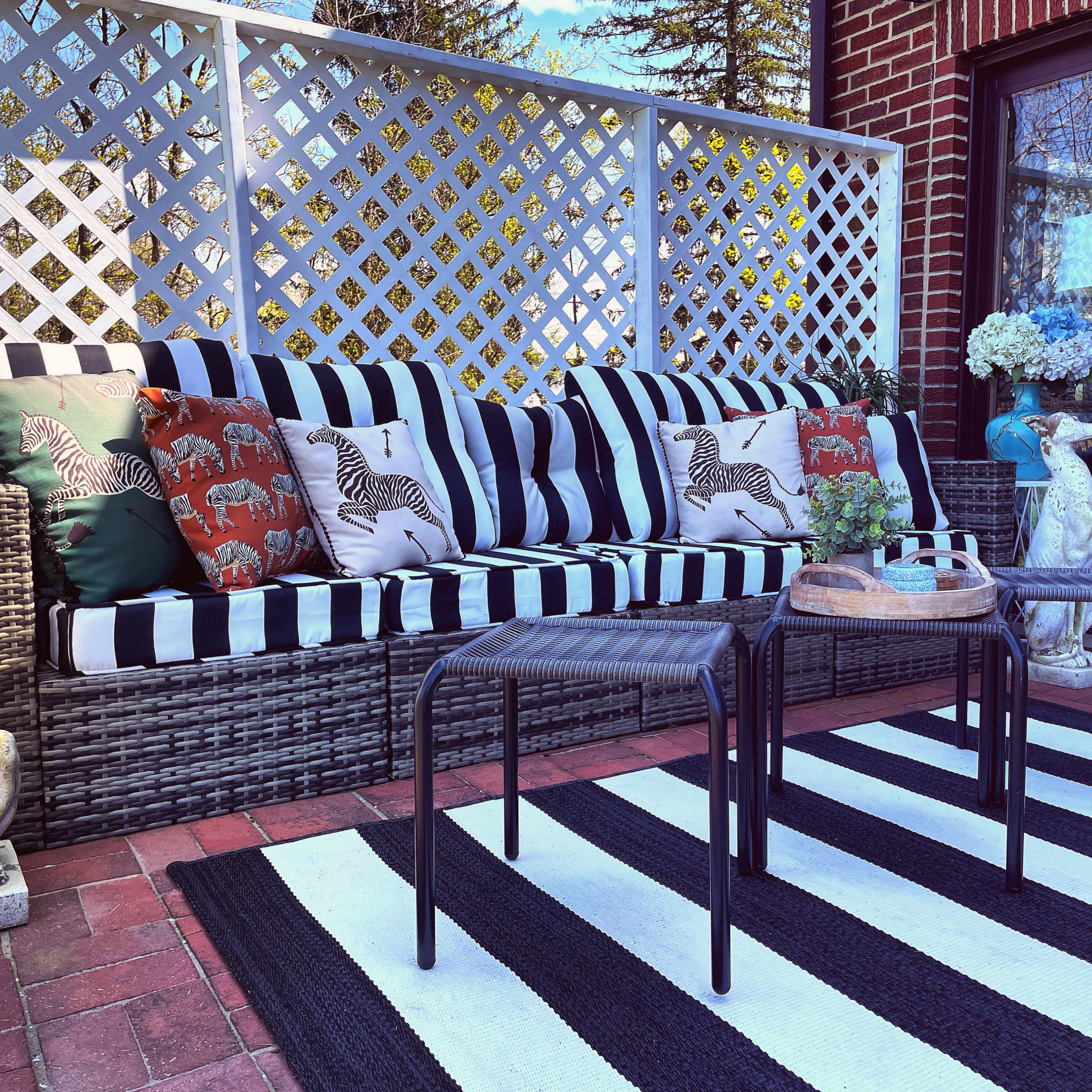 A patio area features a striped sofa adorned with decorative pillows, accompanied by three black side tables on a striped rug and surrounded by a lattice fence.