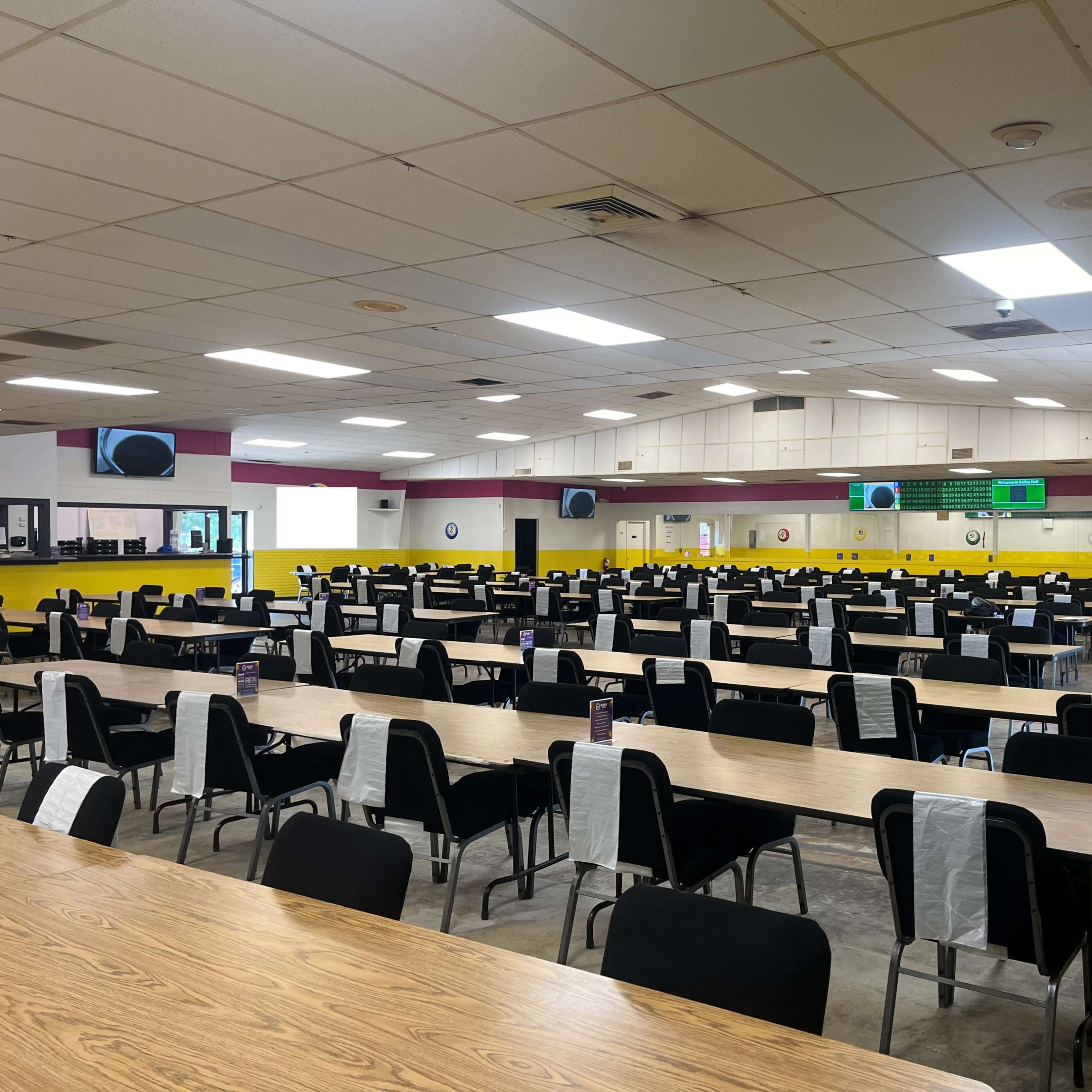 A large cafeteria with numerous empty tables and chairs arranged in rows, illuminated by overhead lights.