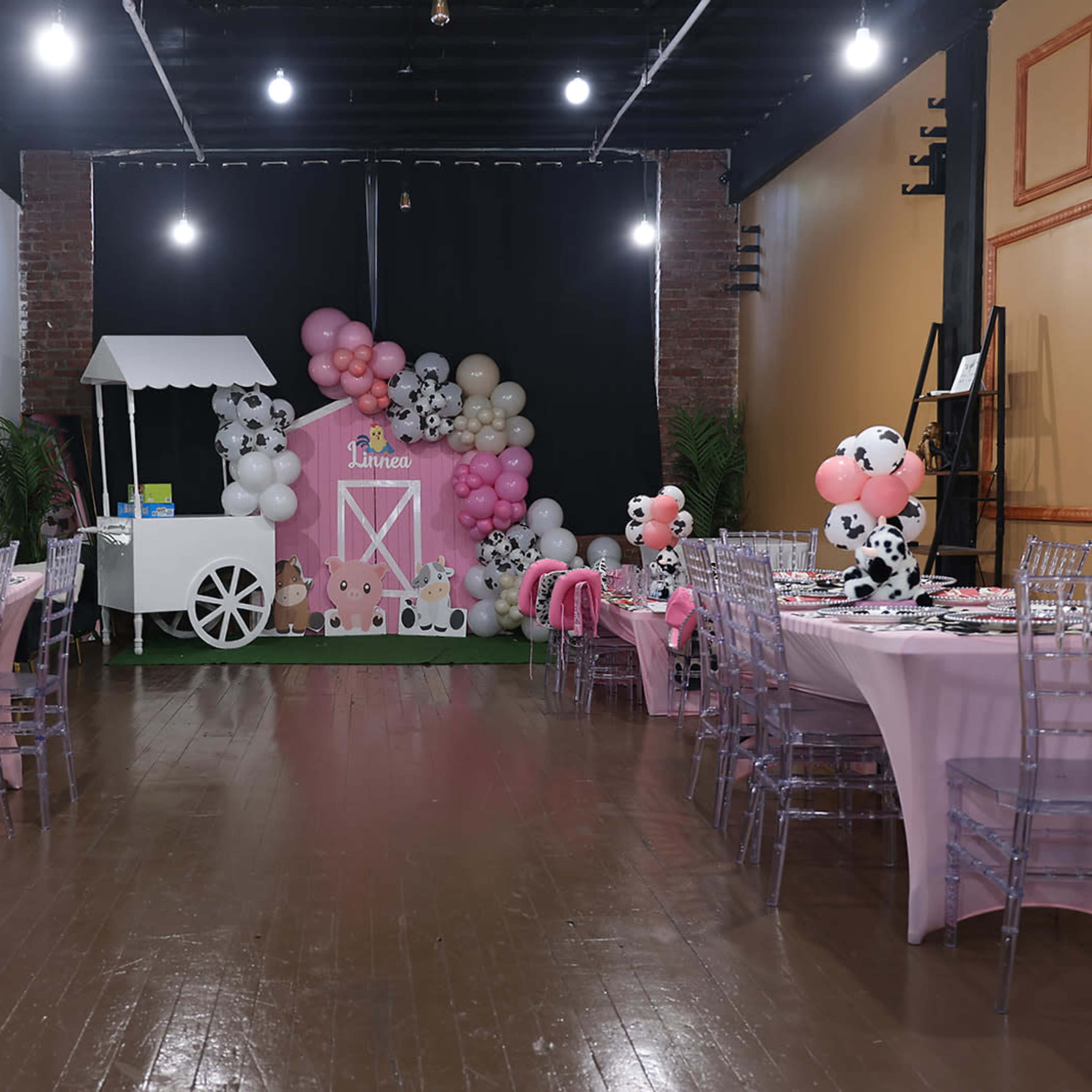 A decorated event space features pink tables with cow-print balloons and a barn-themed backdrop.