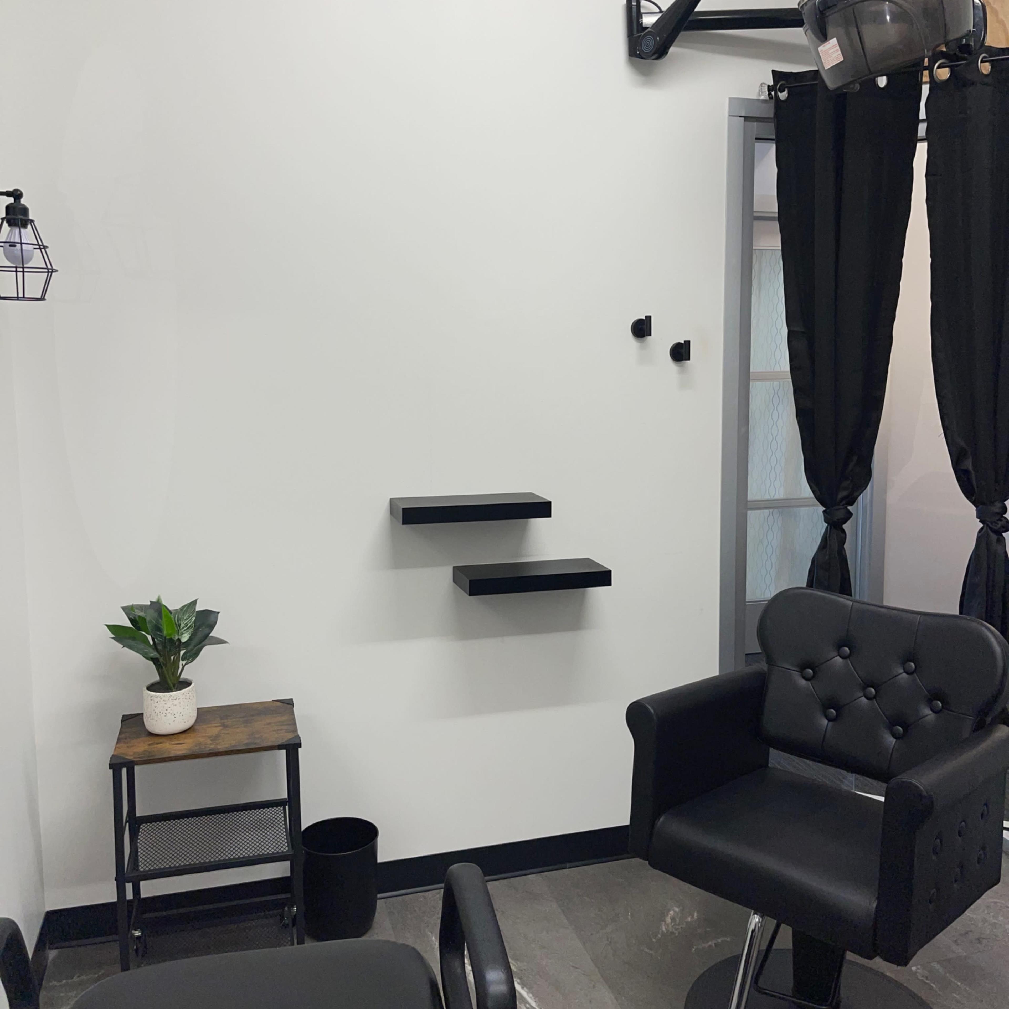 A minimalist hair salon interior featuring a black salon chair, a plant on a small table, and a hooded hair dryer mounted on the wall.