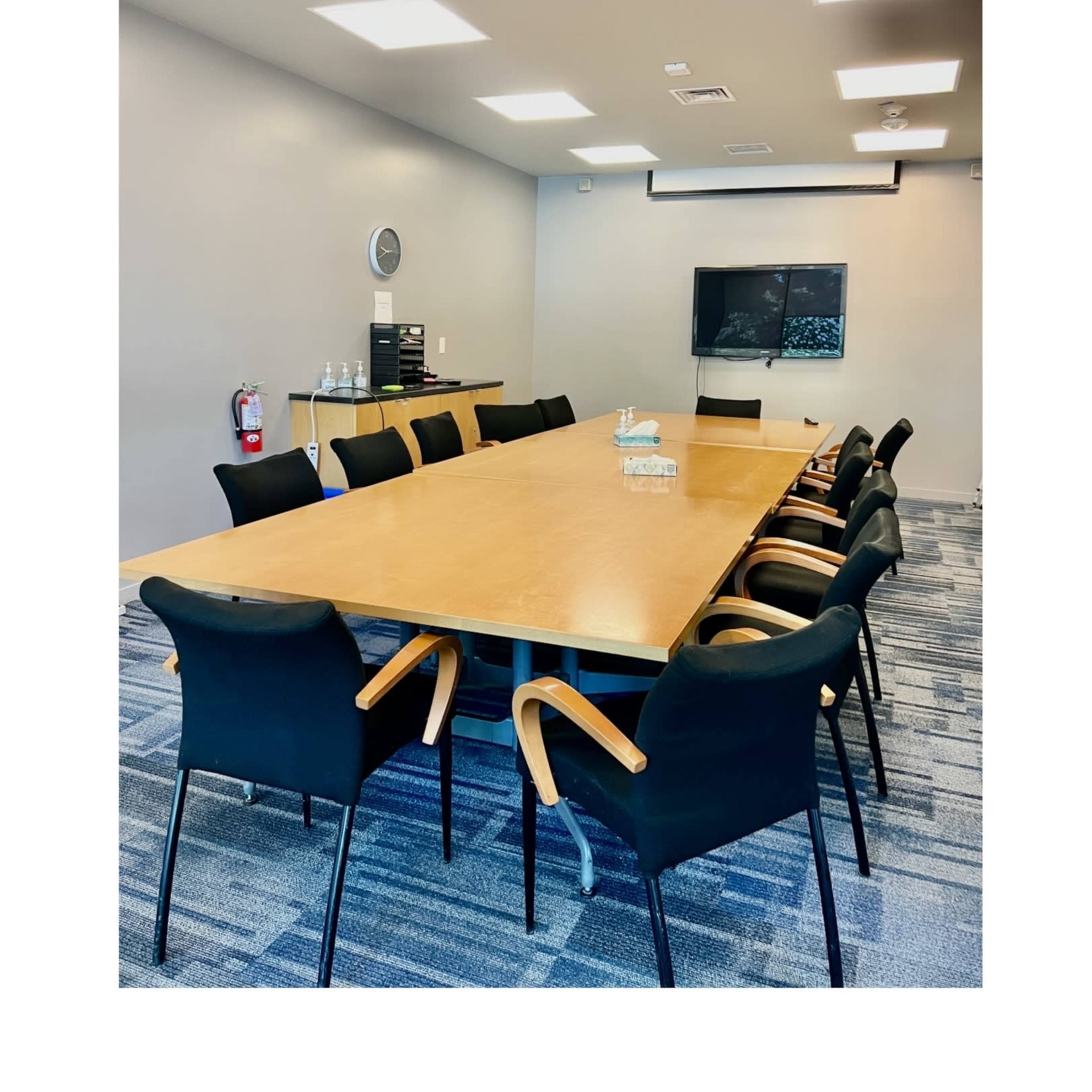 A conference room features a large wooden table surrounded by black chairs, with a wall-mounted television and equipment in the background.