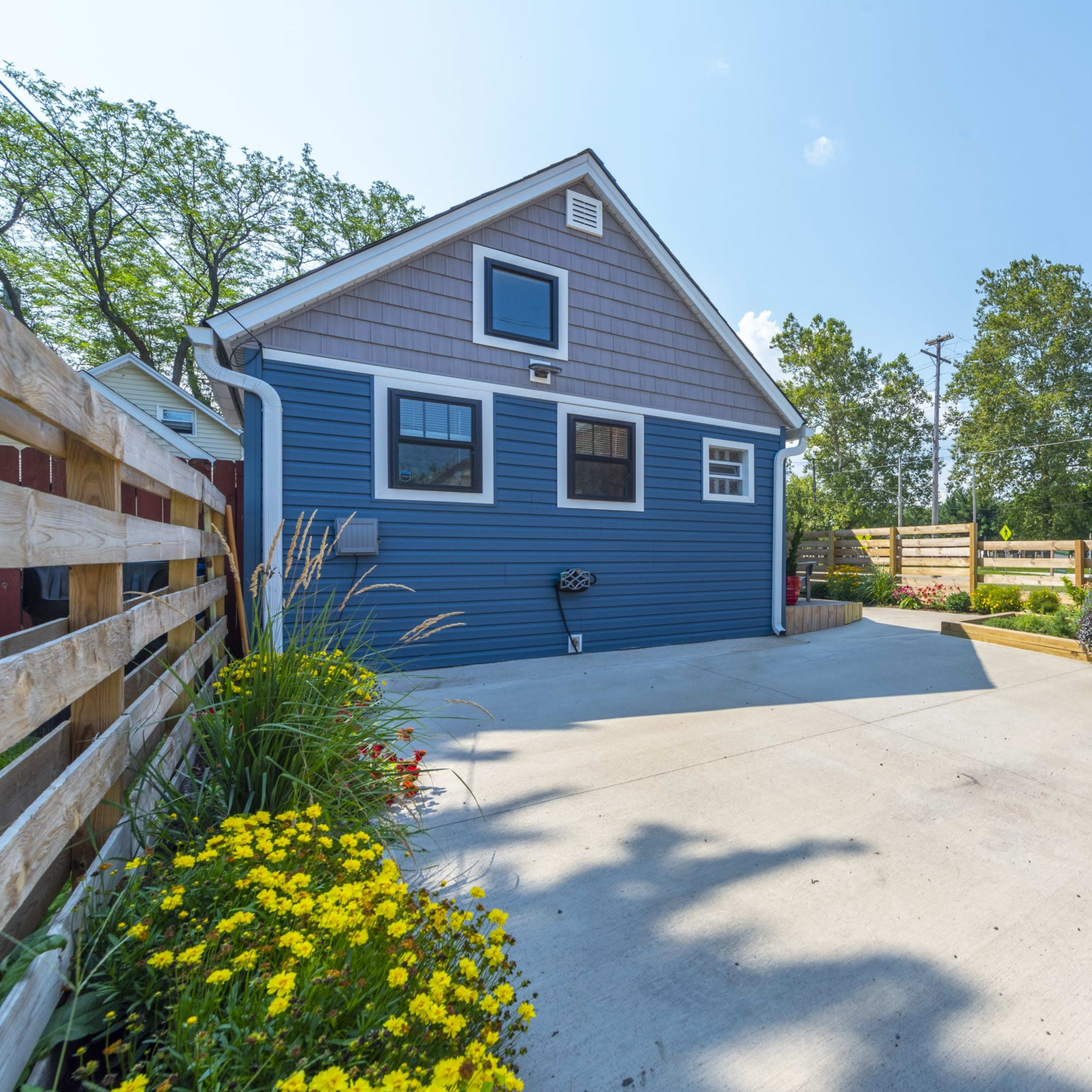 A blue house with a gray roof, a concrete driveway, and a landscape featuring flowers and shrubs, surrounded by a wooden fence.