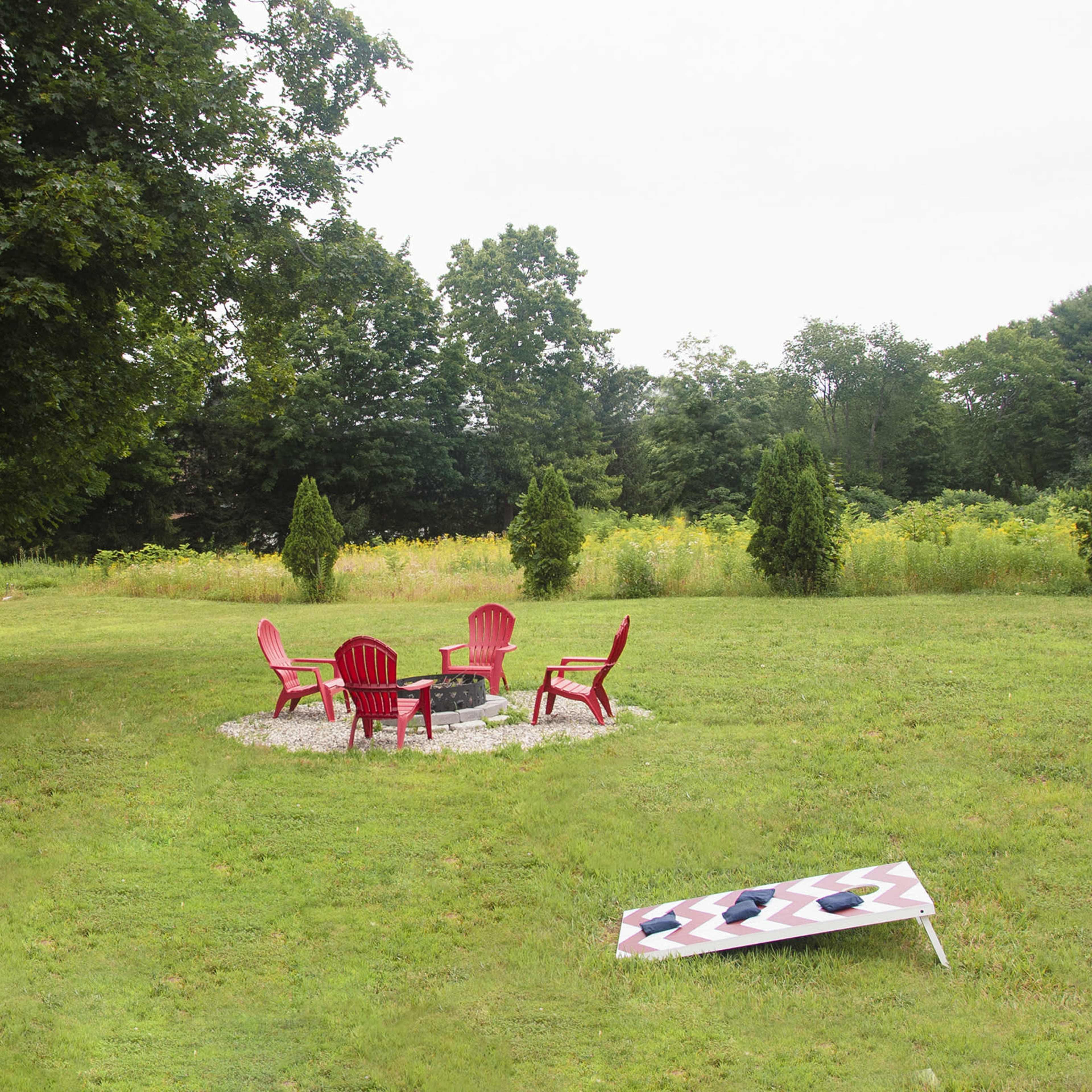 A grassy yard with a circular seating area surrounded by four red chairs and a fire pit, flanked by two bean bag toss boards.
