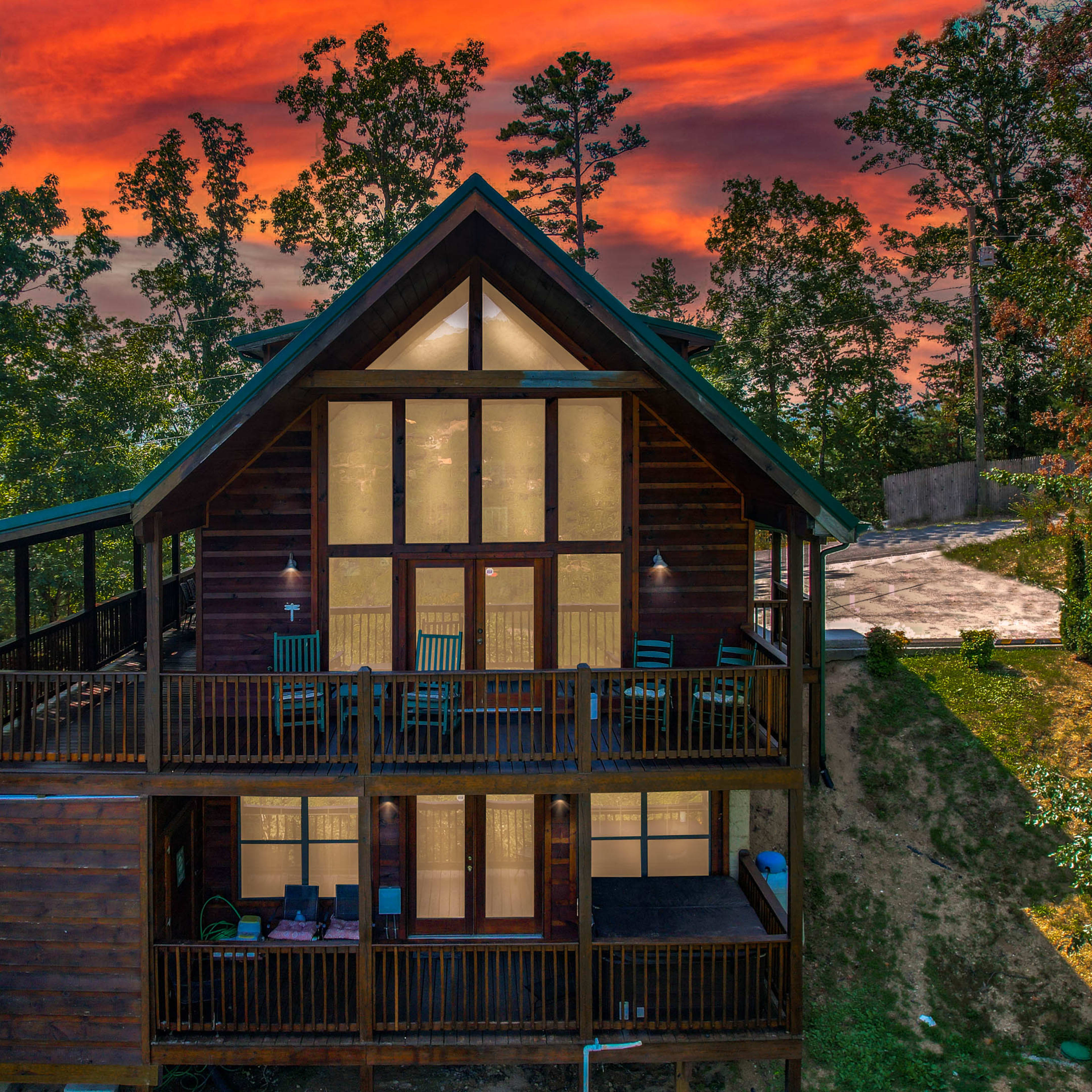 A wooden cabin with large glass windows and multiple balconies is set against a backdrop of a vibrant sunset and surrounded by trees.