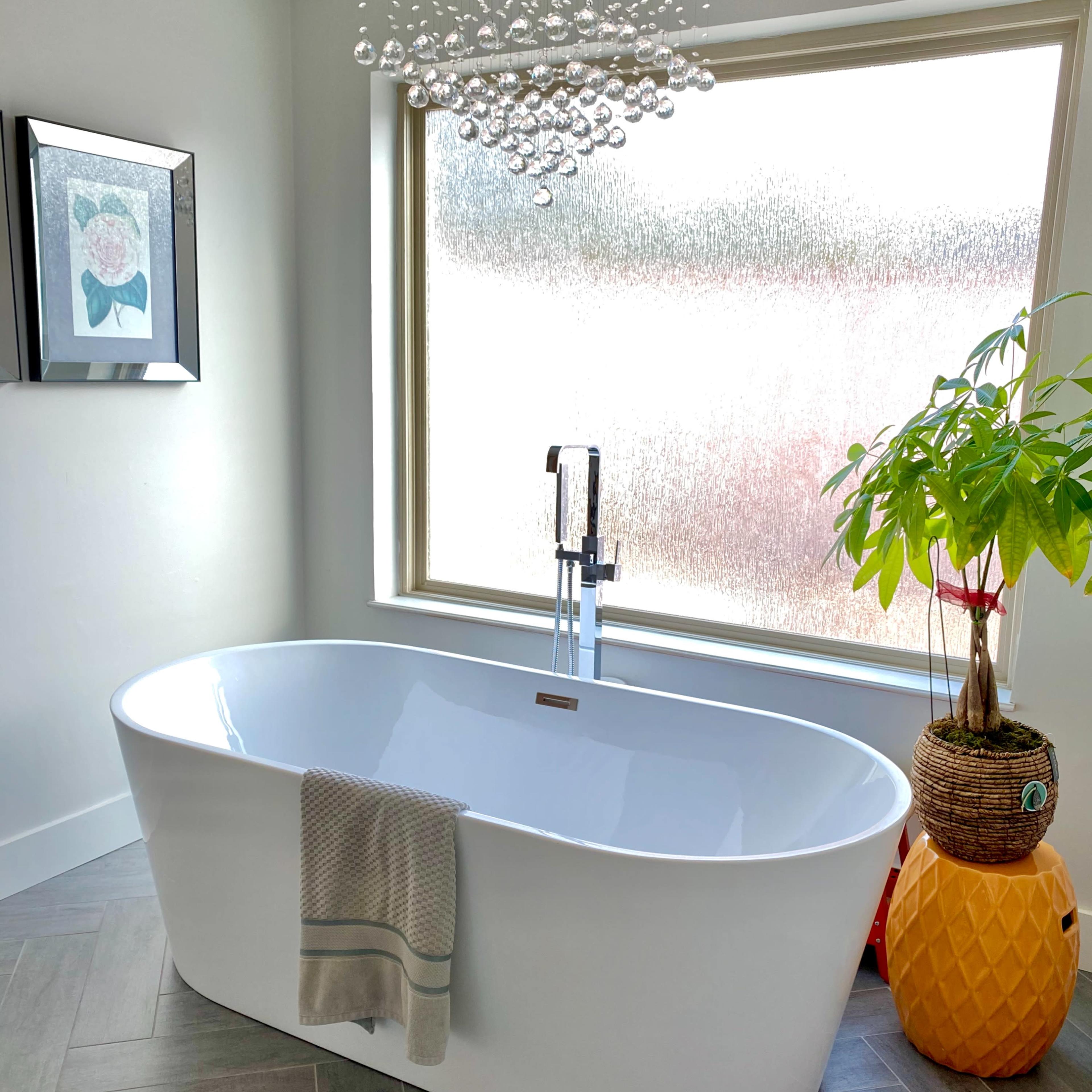 A white freestanding bathtub in a modern bathroom beside a large window with frosted glass, adorned with a plant and decorative elements.