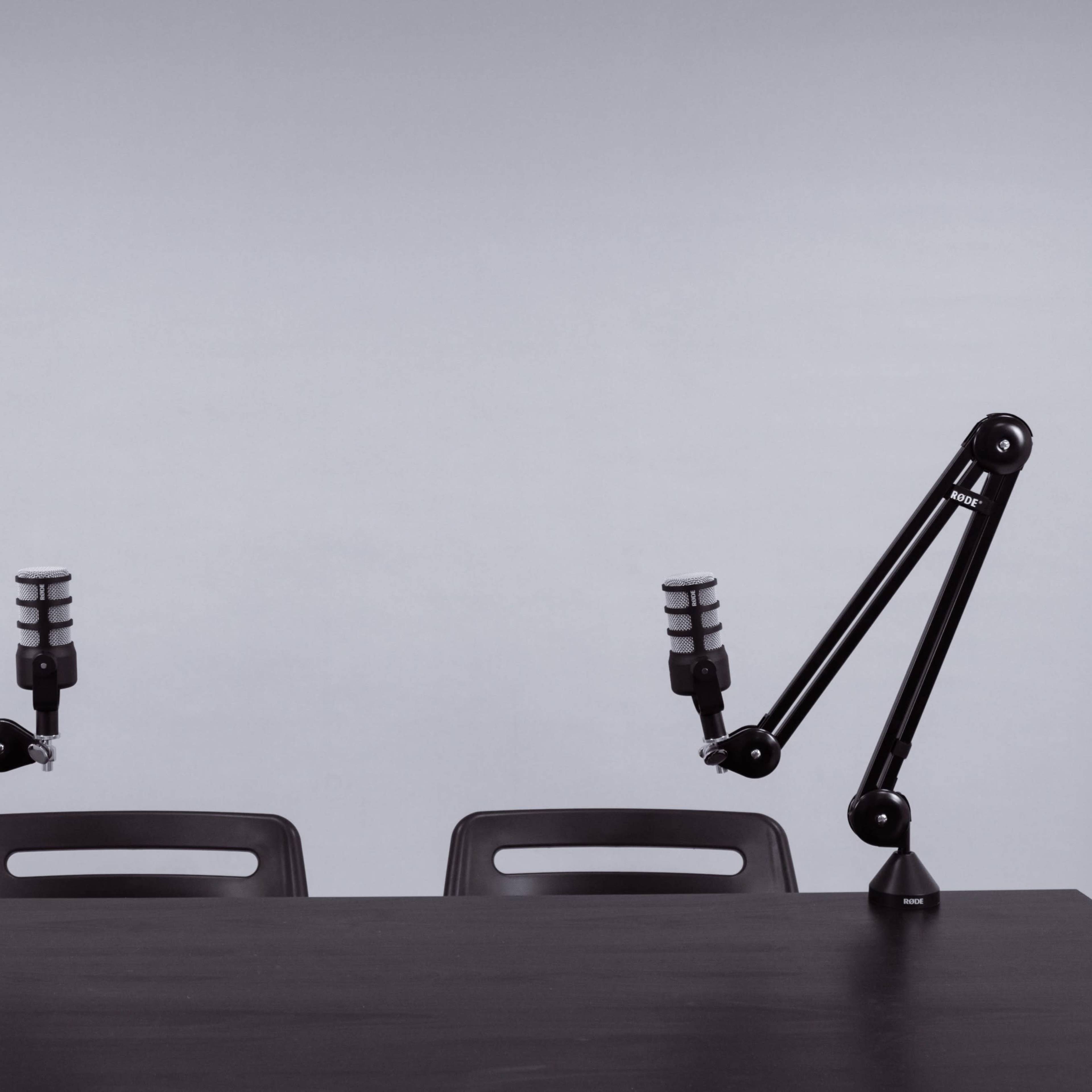 A minimalist podcasting setup features two microphones mounted on adjustable arms positioned over a simple black table with two empty chairs.