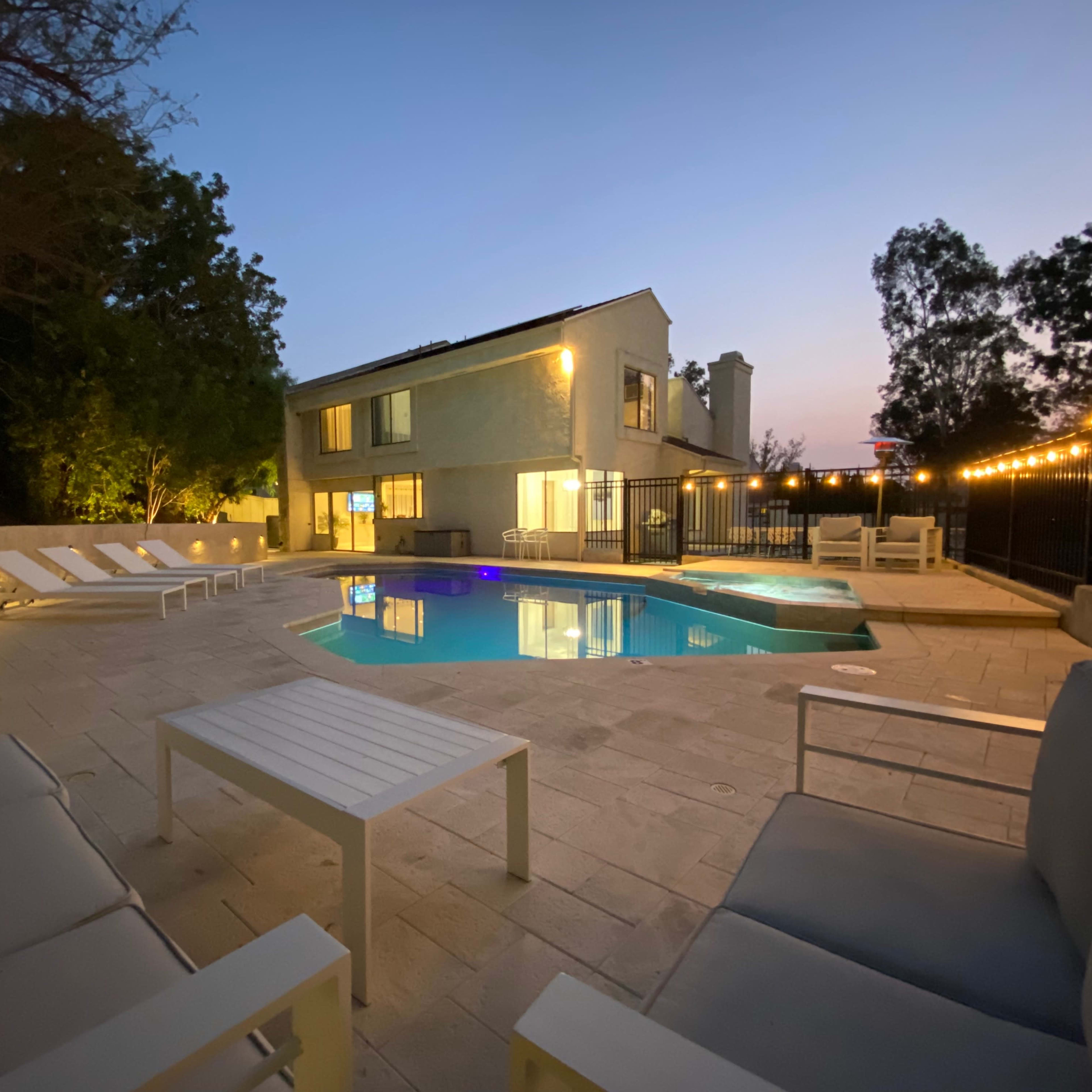 A modern two-story house overlooks a swimming pool surrounded by lounge chairs and illuminated by string lights during twilight.