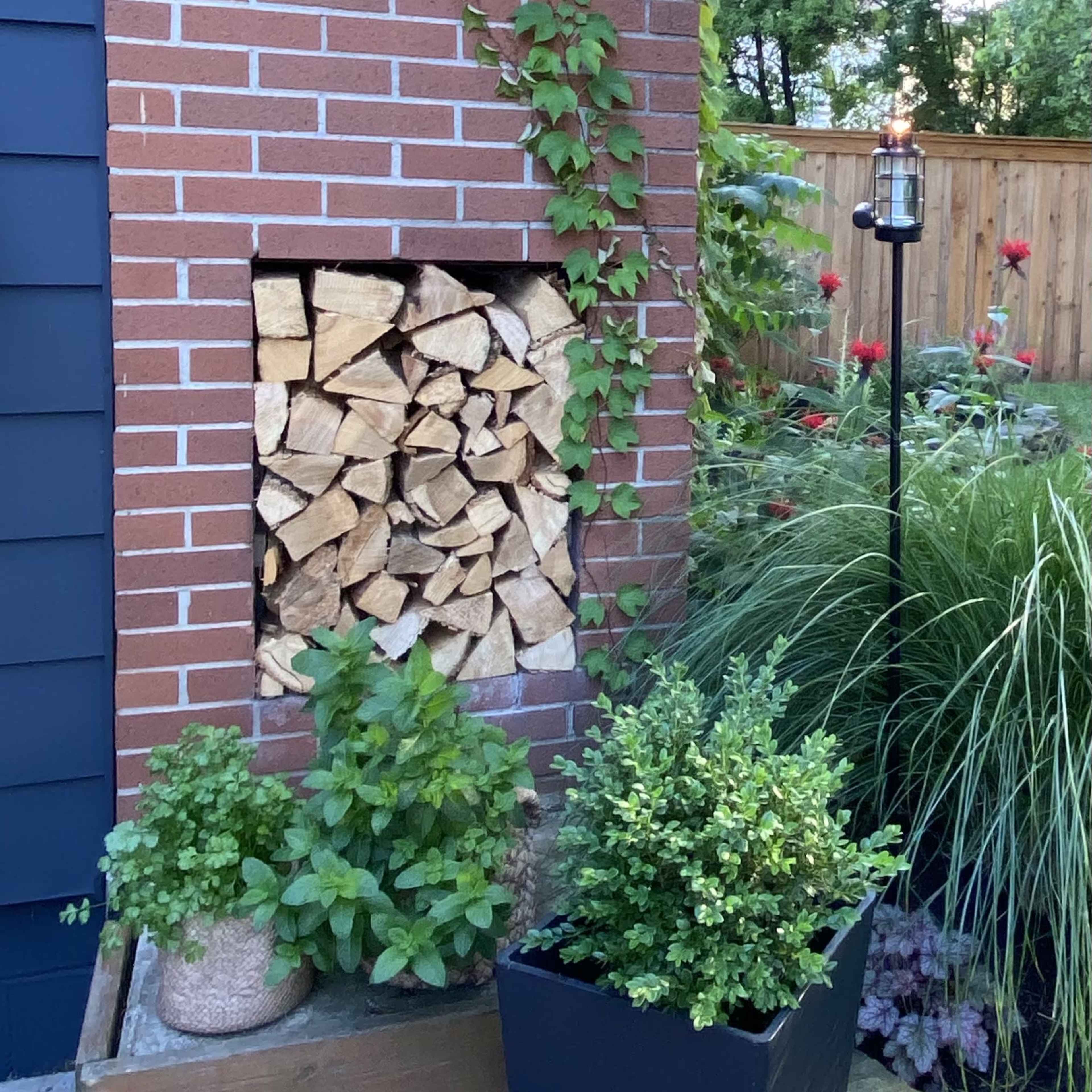 A brick wall features a sun decoration and a stacked wood pile, surrounded by various potted plants and greenery.