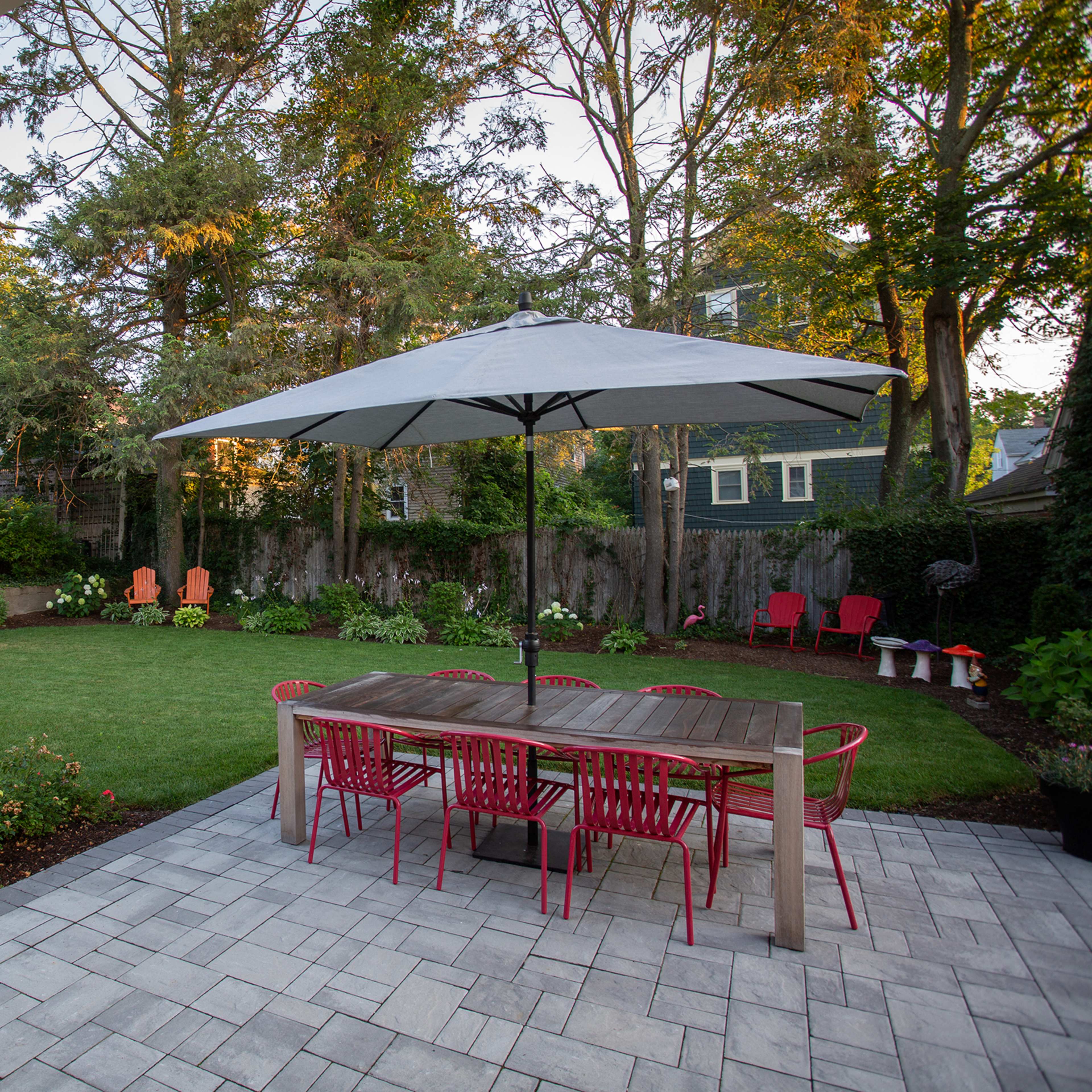A backyard features a wooden dining table with red chairs under a large umbrella, surrounded by green lawn and decorative seating areas.