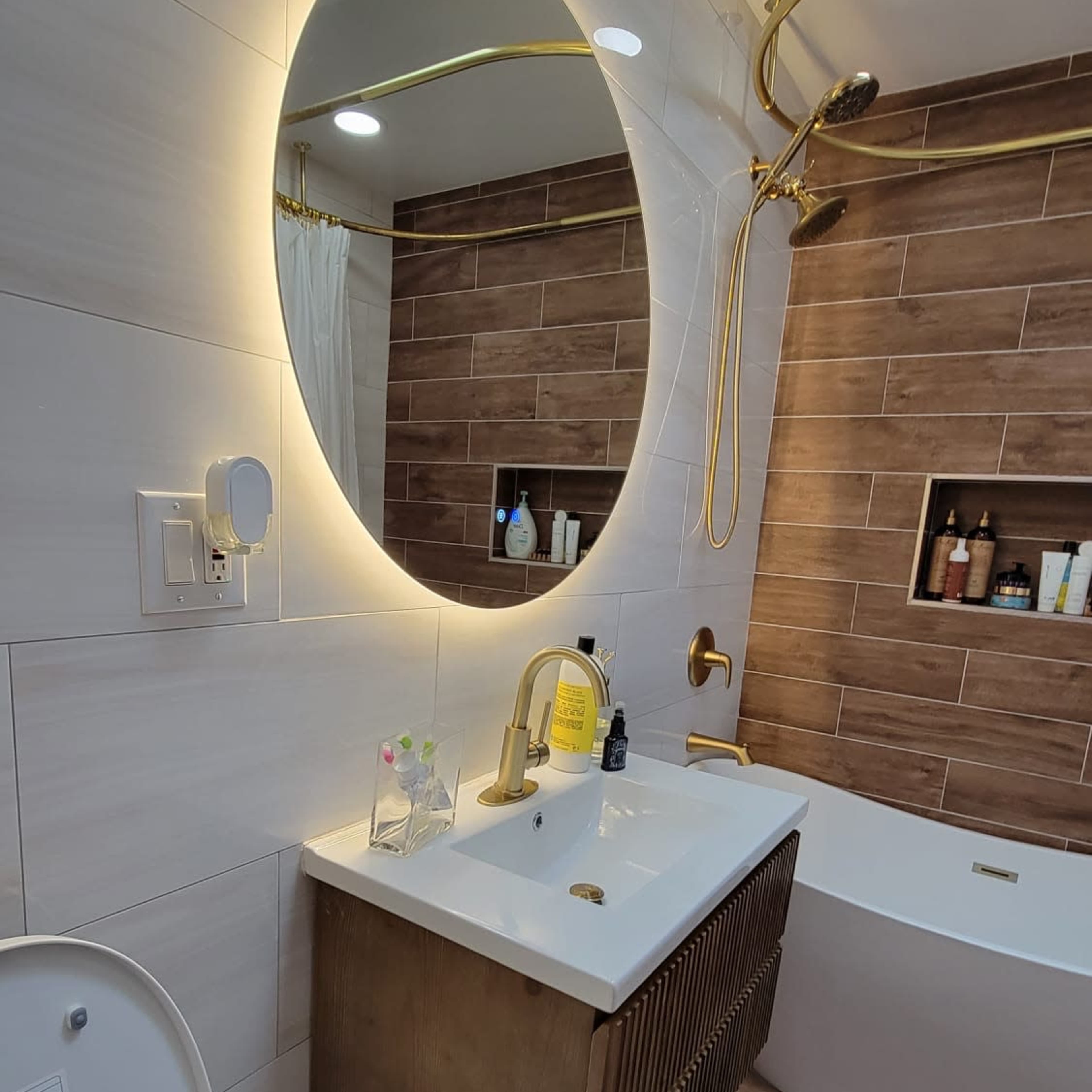 A modern bathroom features a round mirror with backlighting, a gold faucet, and wooden accents on the vanity, with a bathtub and a towel rack in the background.