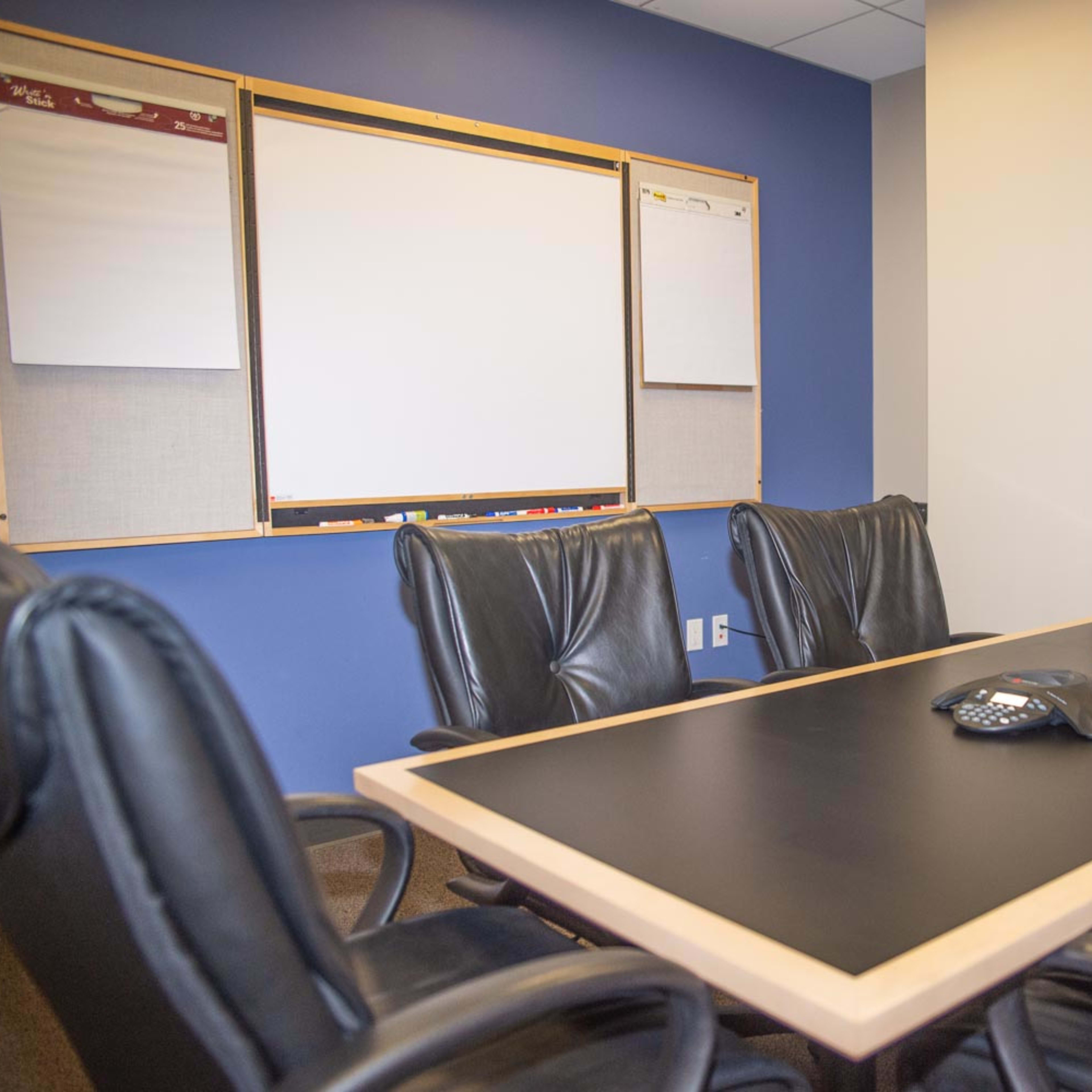 A conference room features a large table surrounded by black leather chairs, with a whiteboard and flip charts on the wall.