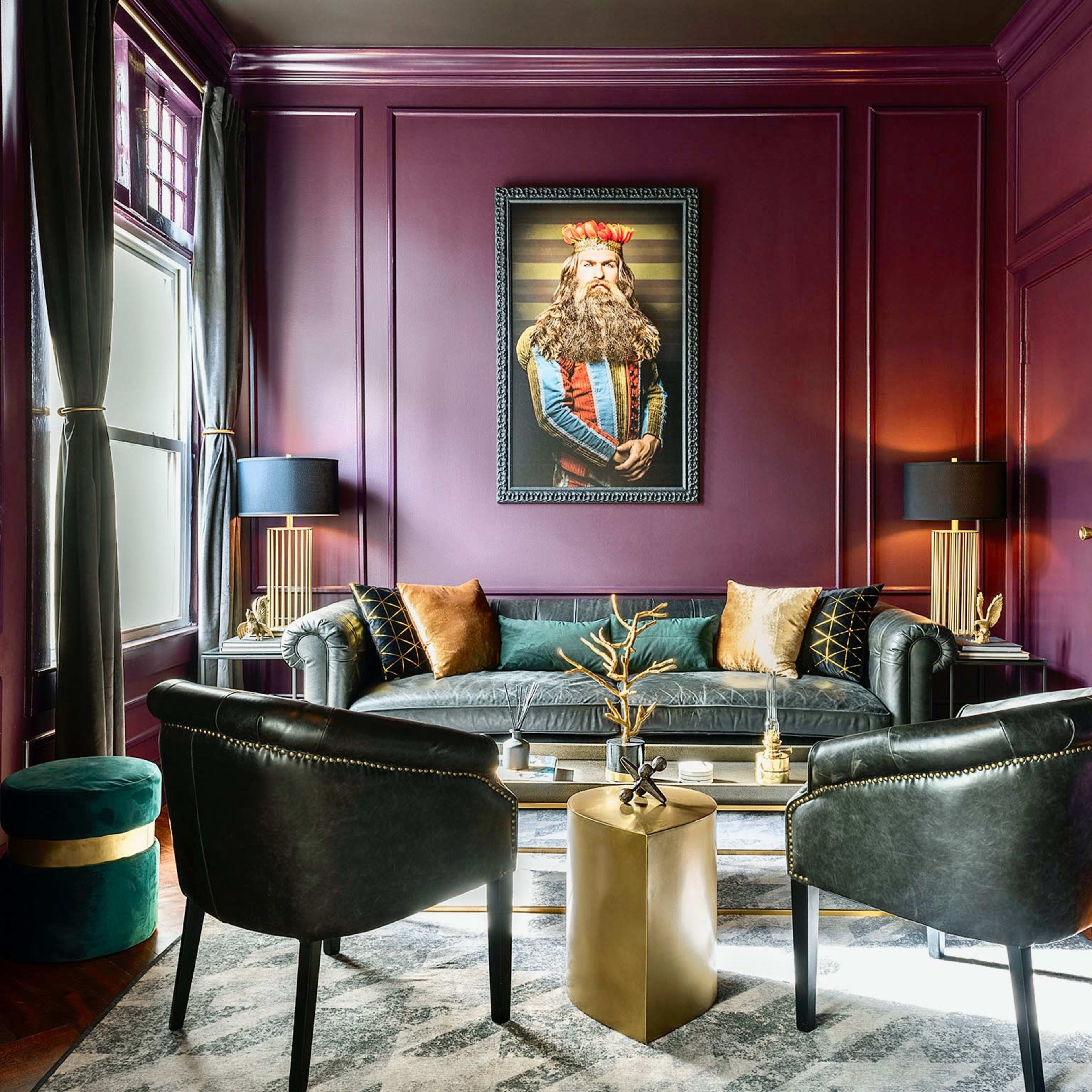 A stylish living room with a deep purple wall, a black leather sofa, two black chairs, and a large portrait of a king hanging above the sofa.