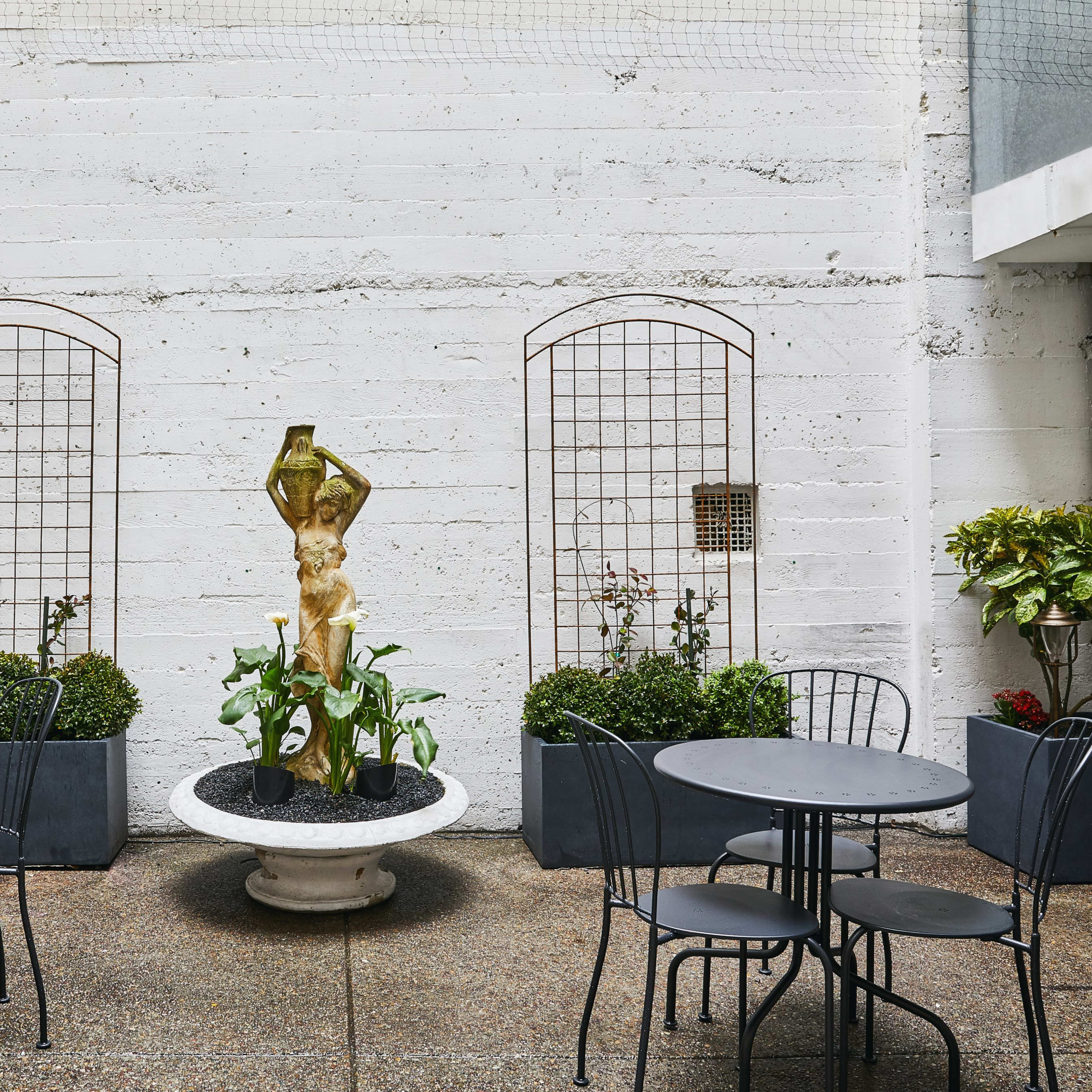 A courtyard with four round, black metal tables and chairs surrounding a central golden statue in a planter, flanked by trellises and greenery against a textured concrete wall.
