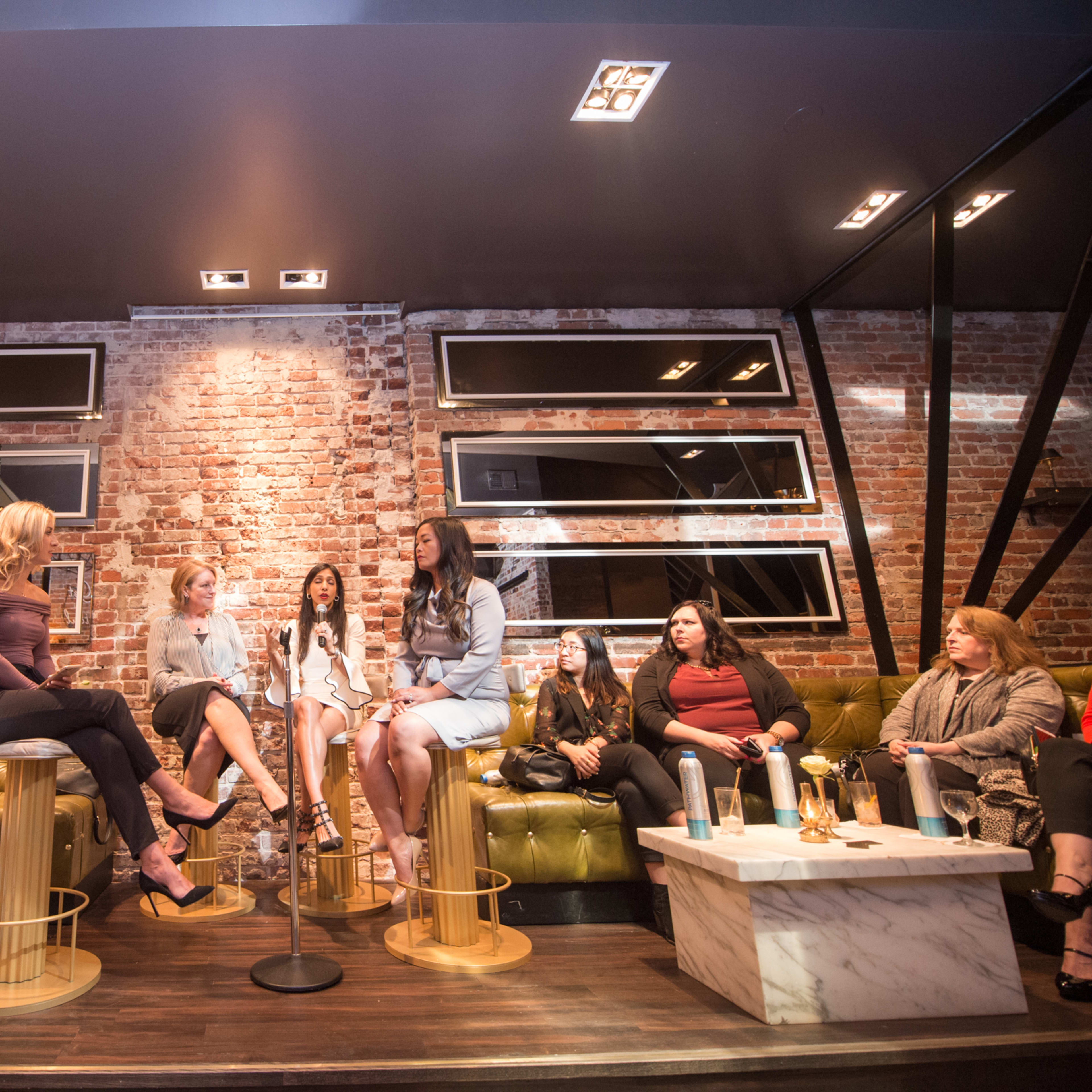 A group of women is seated in a lounge, engaged in conversation while sitting on bar stools and a plush sofa against a brick wall.