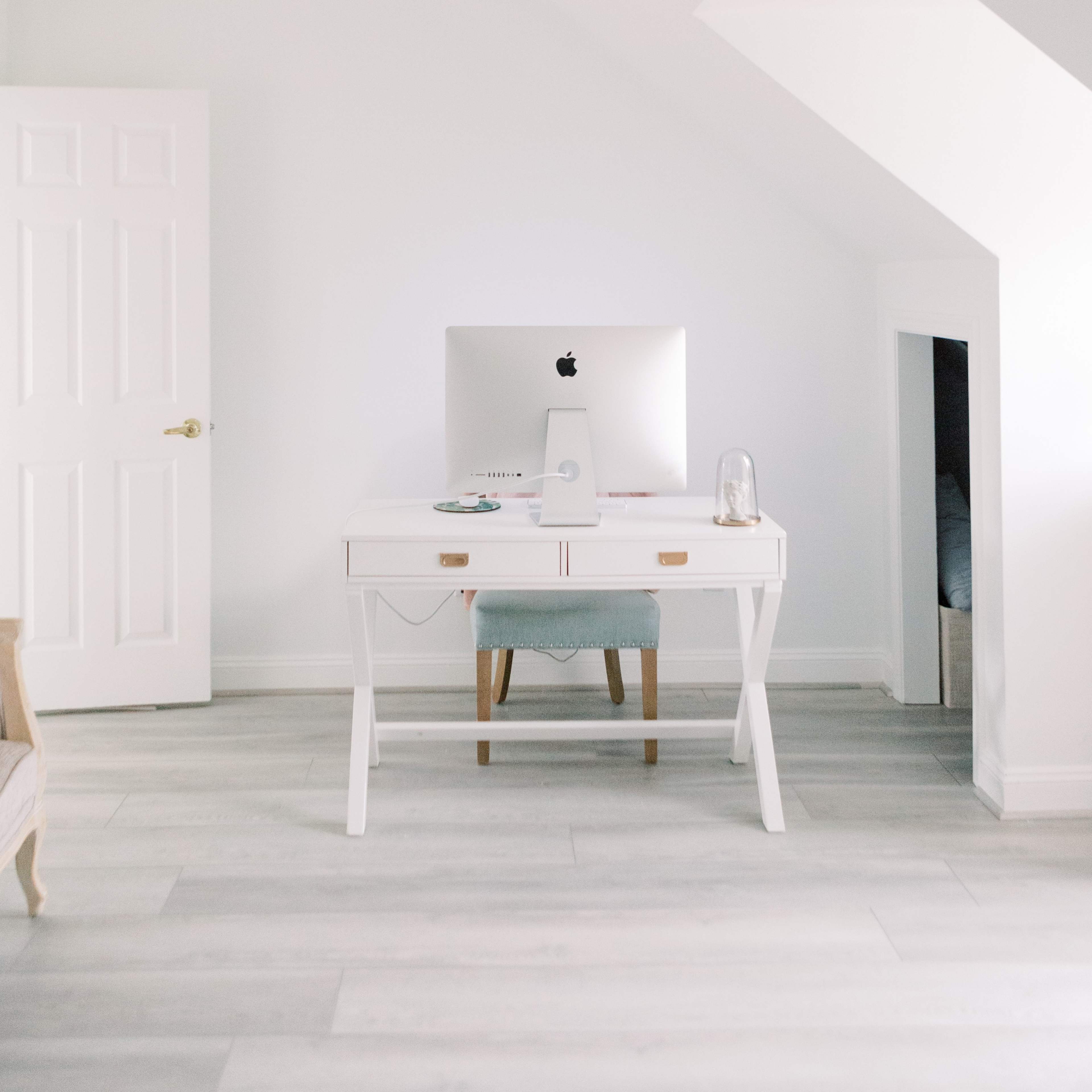 A white desk with a computer sits in a minimalist room with light-colored wood flooring and a partially open door.