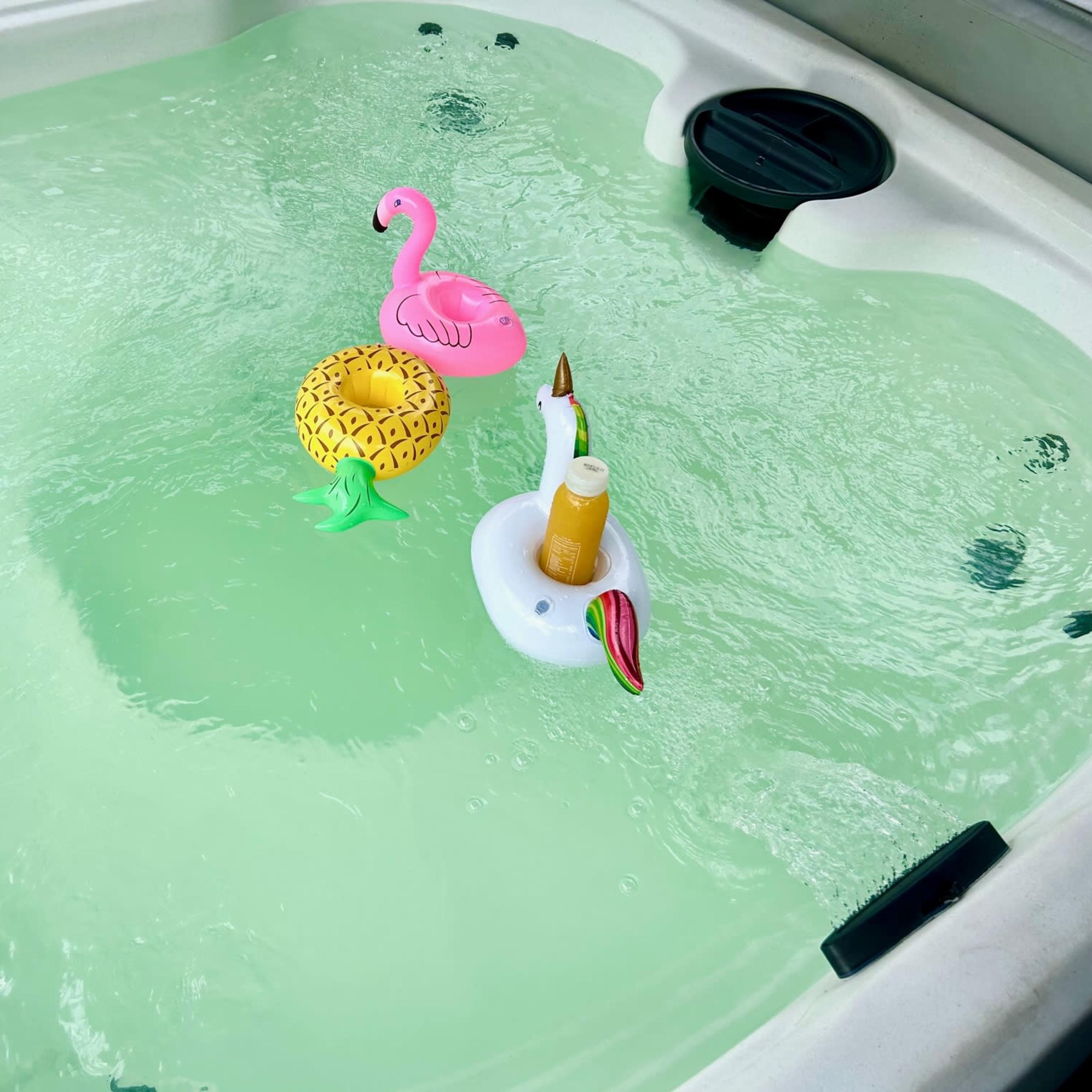 A hot tub filled with green-tinted water contains a pink flamingo float, a pineapple float, and a white float holding a bottle of sunscreen.