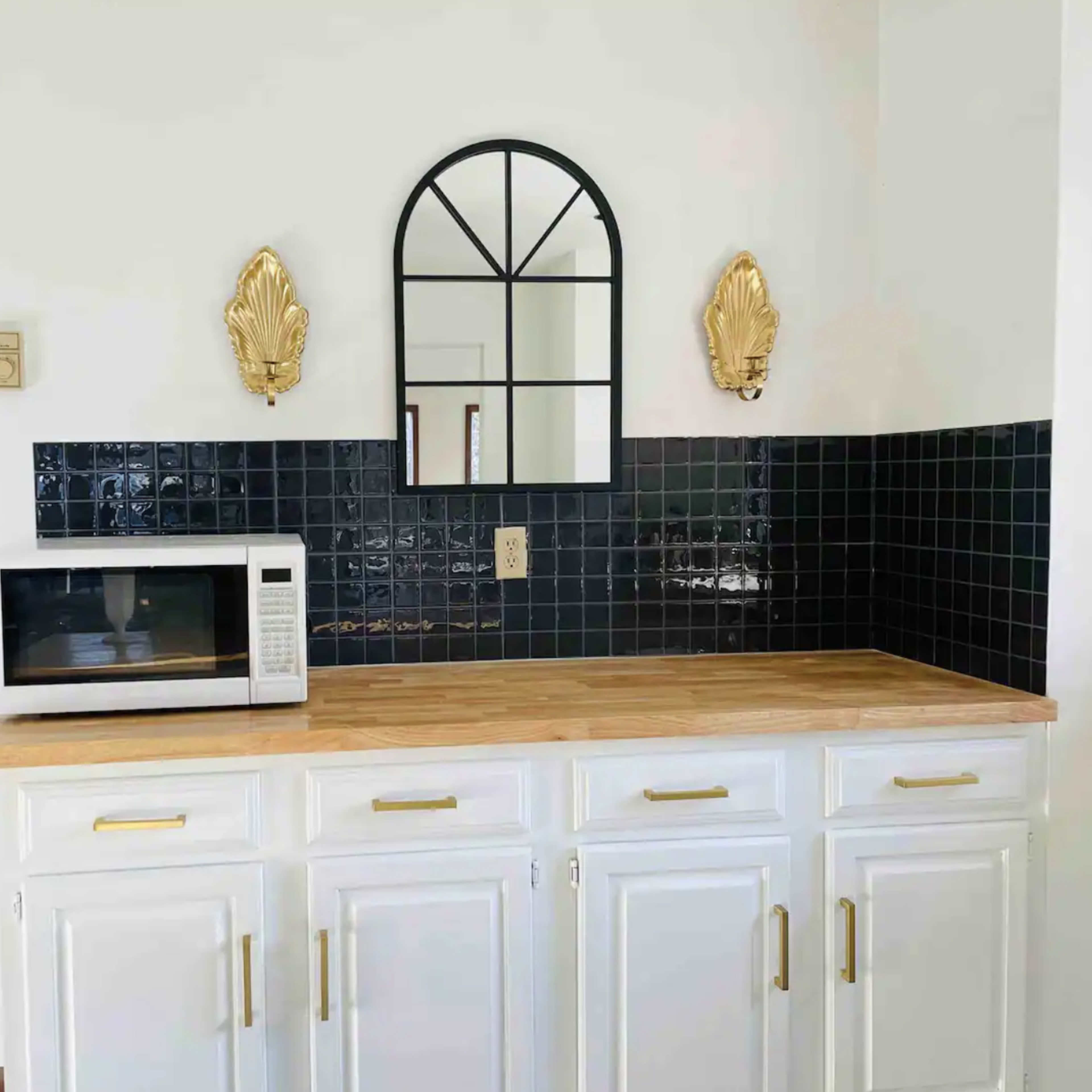 A kitchen countertop with a white microwave, a wooden surface, a decorative mirror, and two wall sconces above dark tiled backsplash.