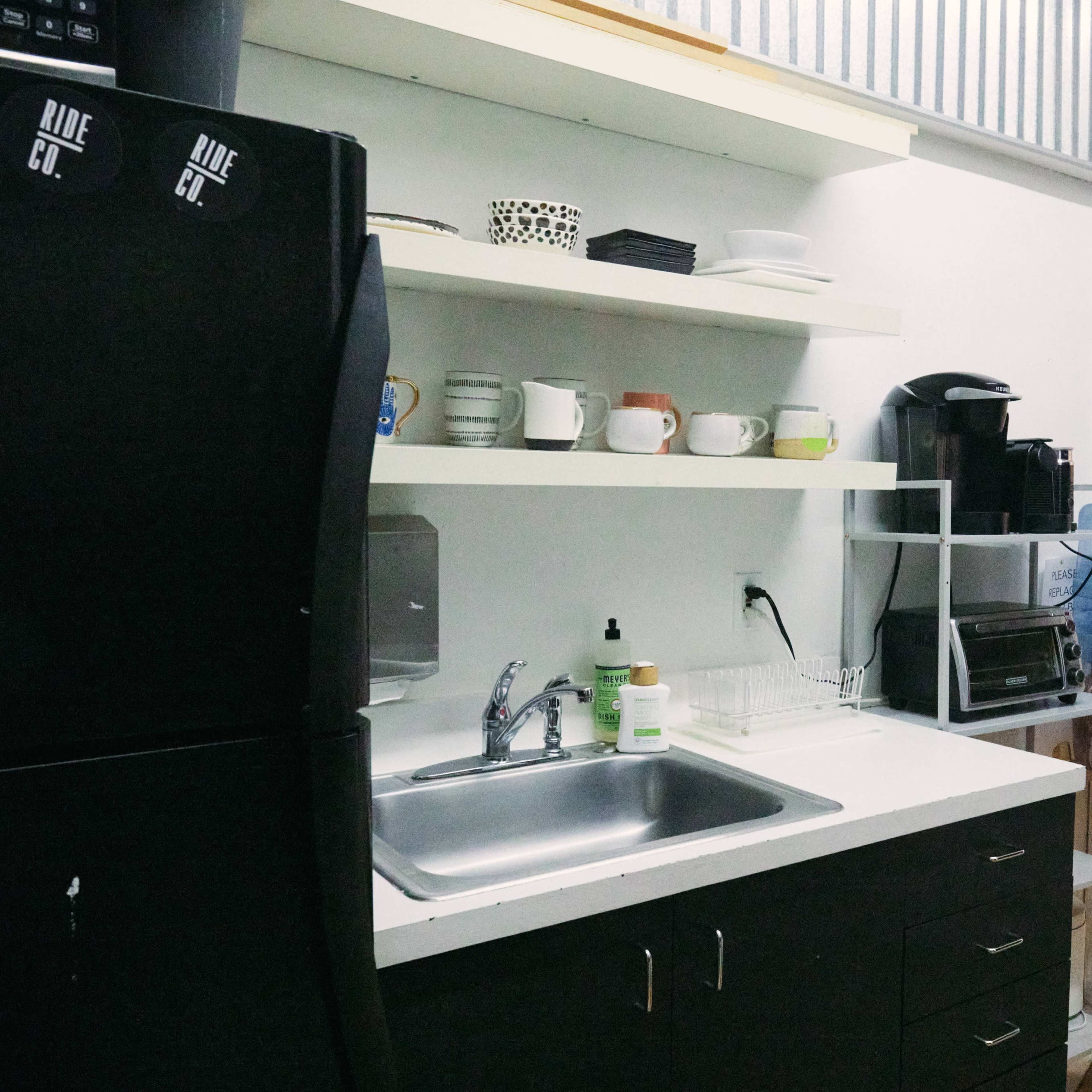 A compact kitchen area with a black refrigerator, a stainless steel sink, shelves holding dishes, and a coffee maker, adjacent to a water cooler and a glass door.
