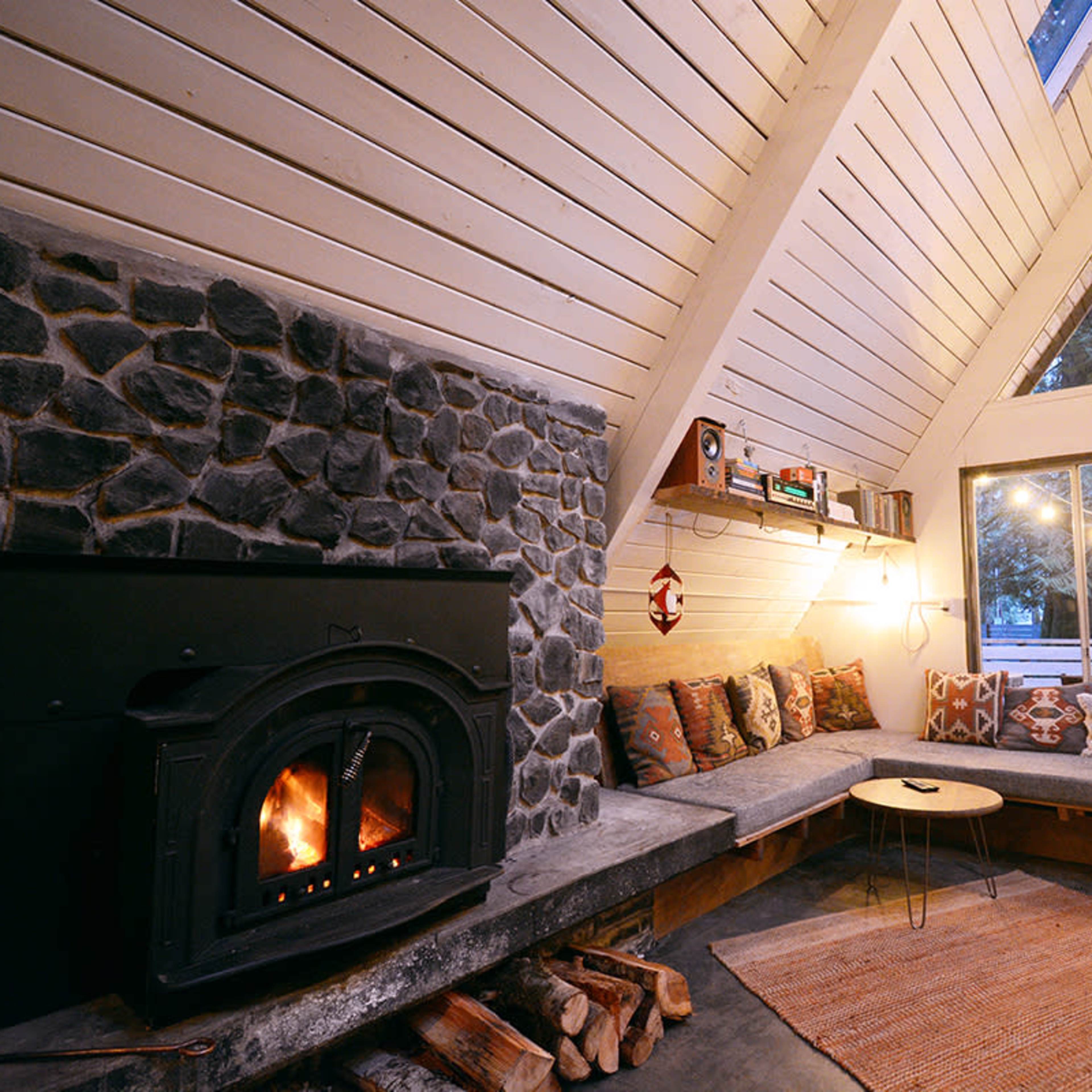 A cozy living room with a stone fireplace, a triangular wooden ceiling, a large window, and a circular coffee table surrounded by a cushioned seating area.