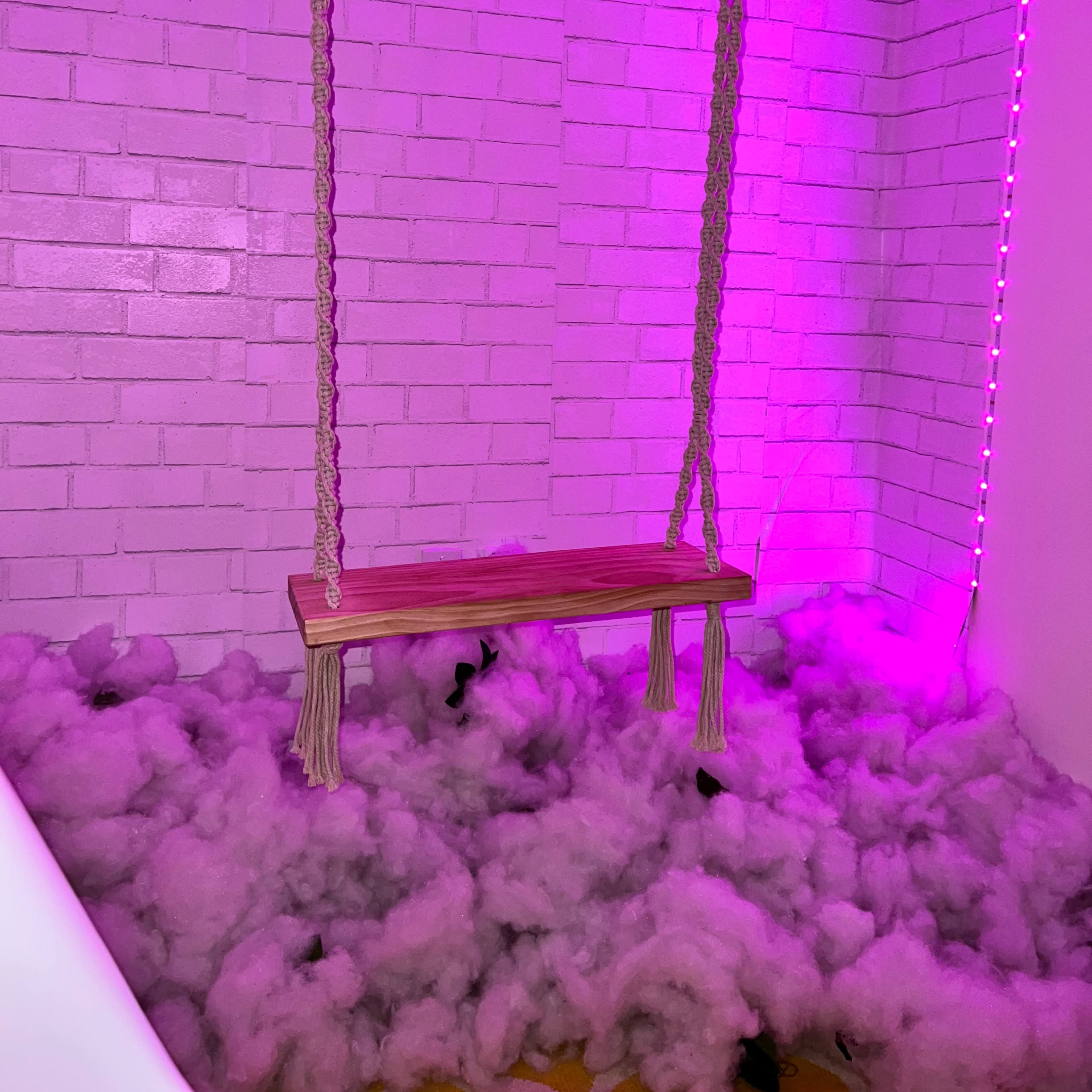 A wooden swing hangs from the ceiling in a corner filled with pink-lit clouds of soft material.