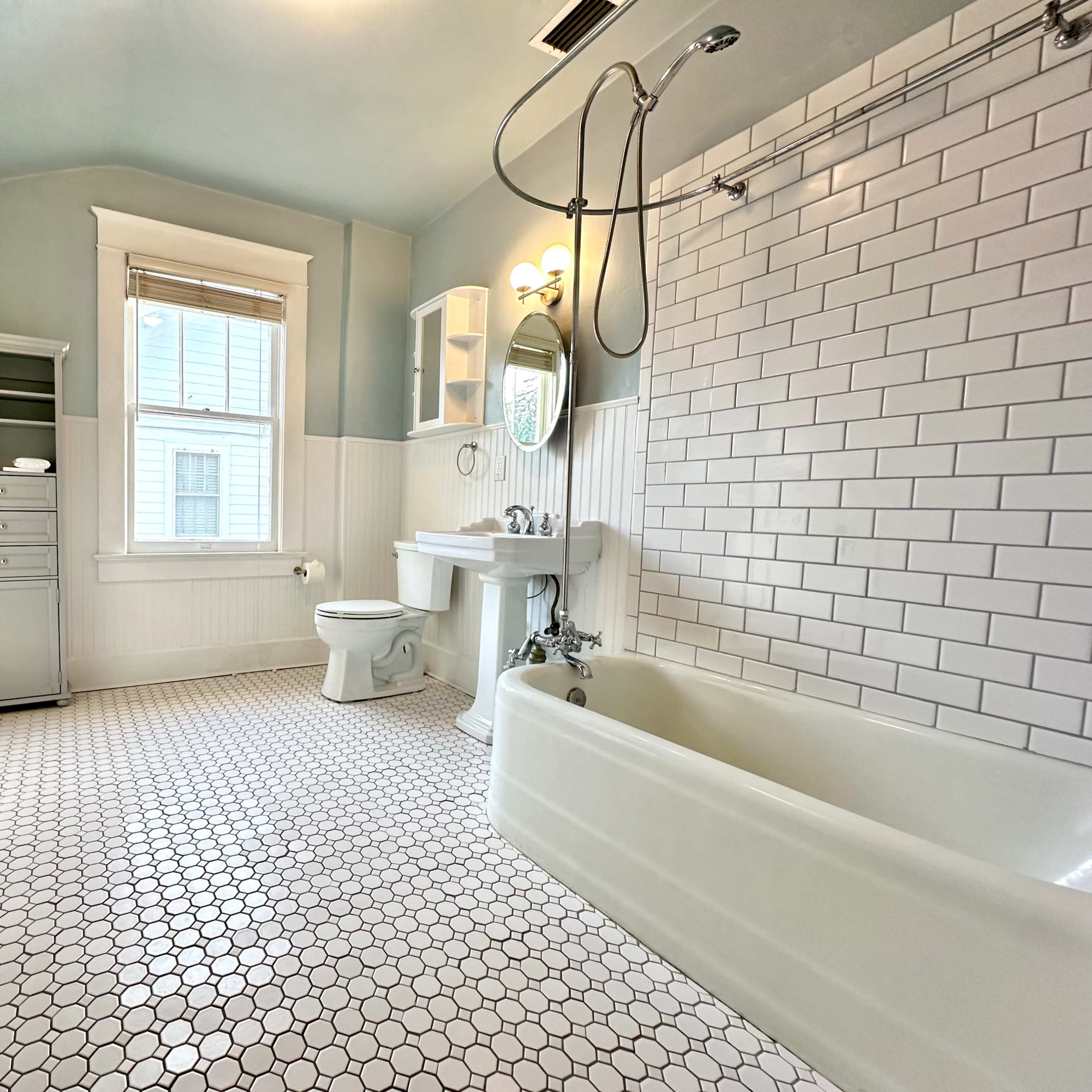A bright bathroom featuring white subway tile walls, a hexagonal tiled floor, a freestanding bathtub, a toilet, and a sink, with natural light coming through a window.