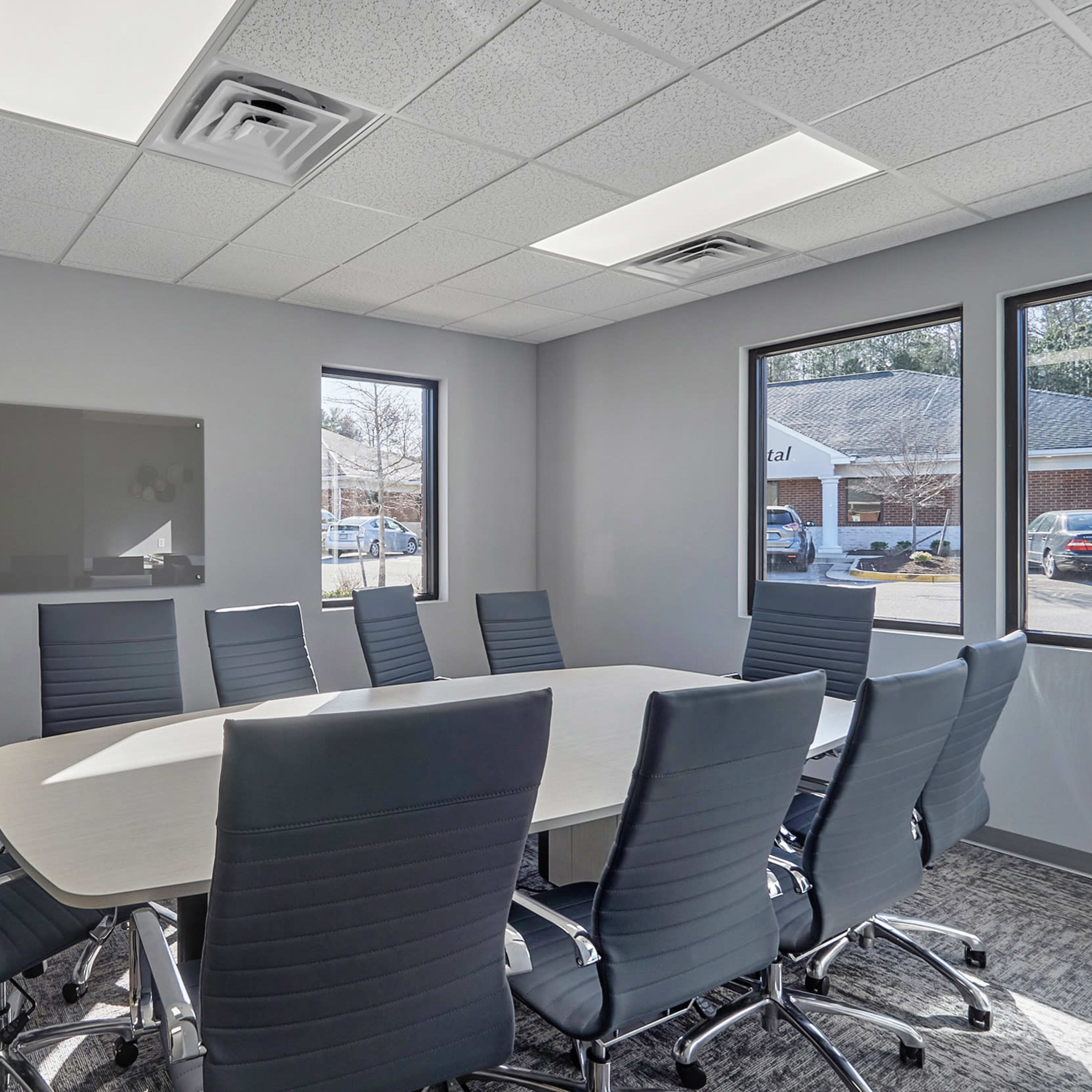 A modern conference room features a large oval table surrounded by ten ergonomic chairs, with windows allowing natural light and a wall-mounted screen.