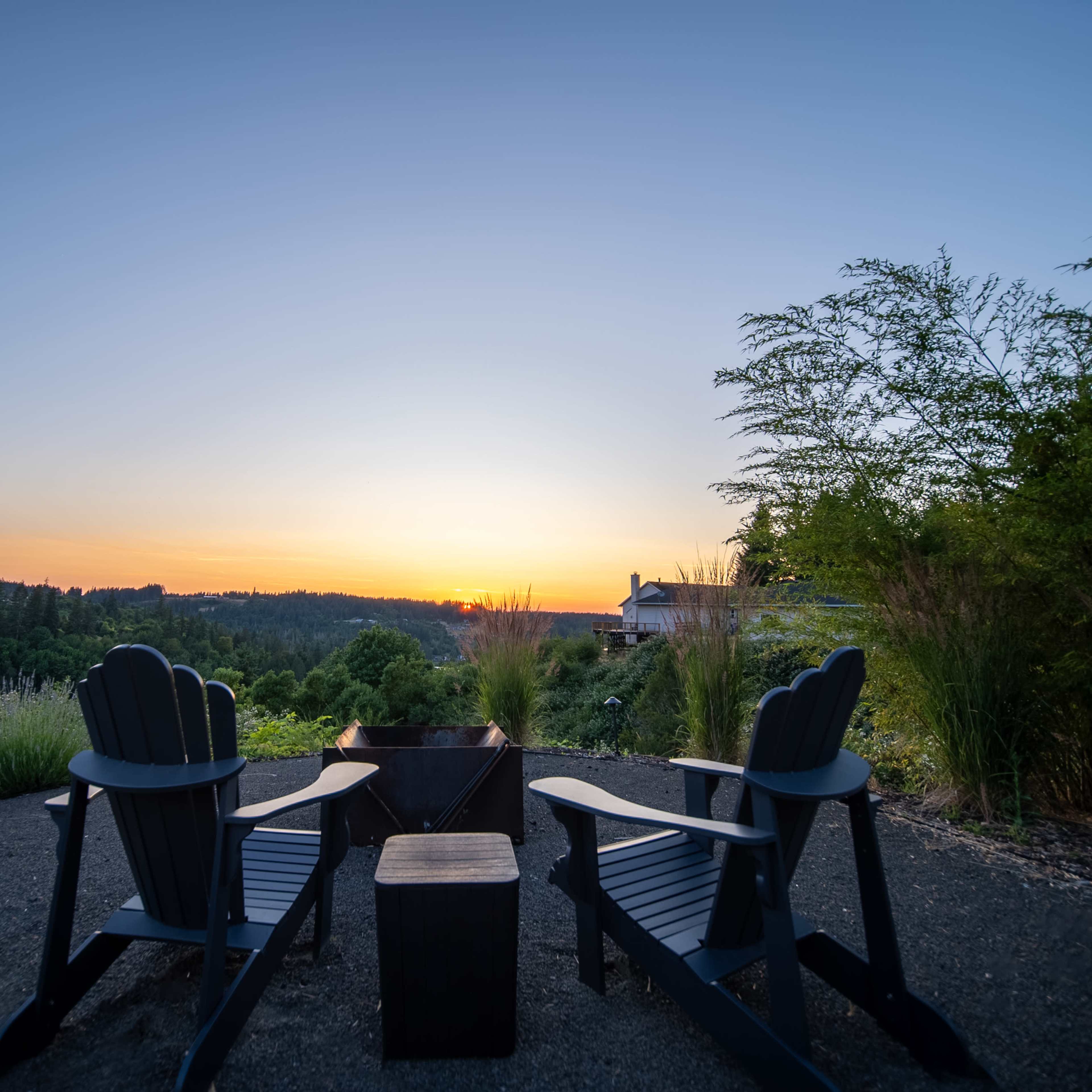 Two black Adirondack chairs facing a fire pit with a sunset view over a lush landscape.