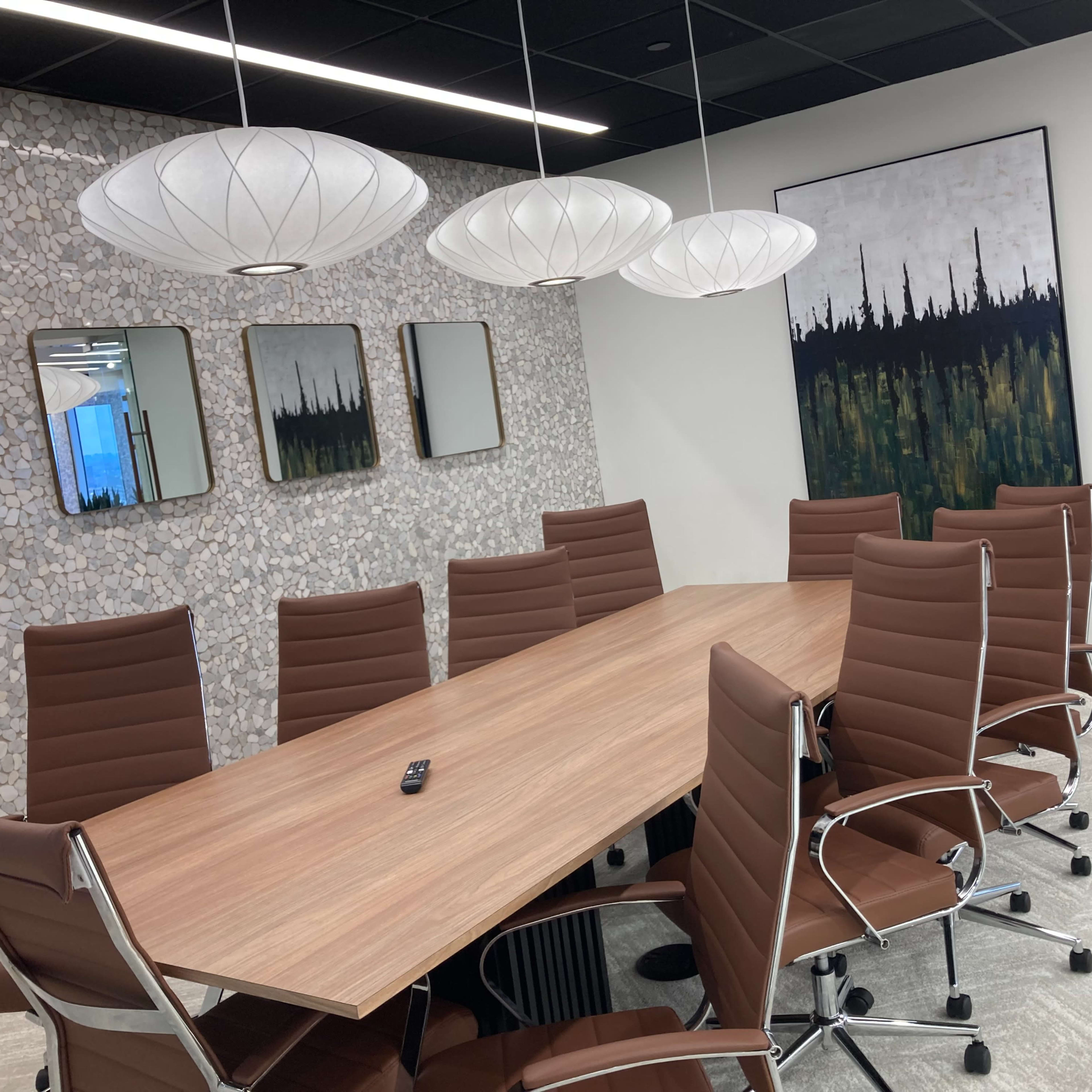 A modern conference room features a long wooden table surrounded by brown leather chairs, with pendant lights overhead and a stone wall accent.