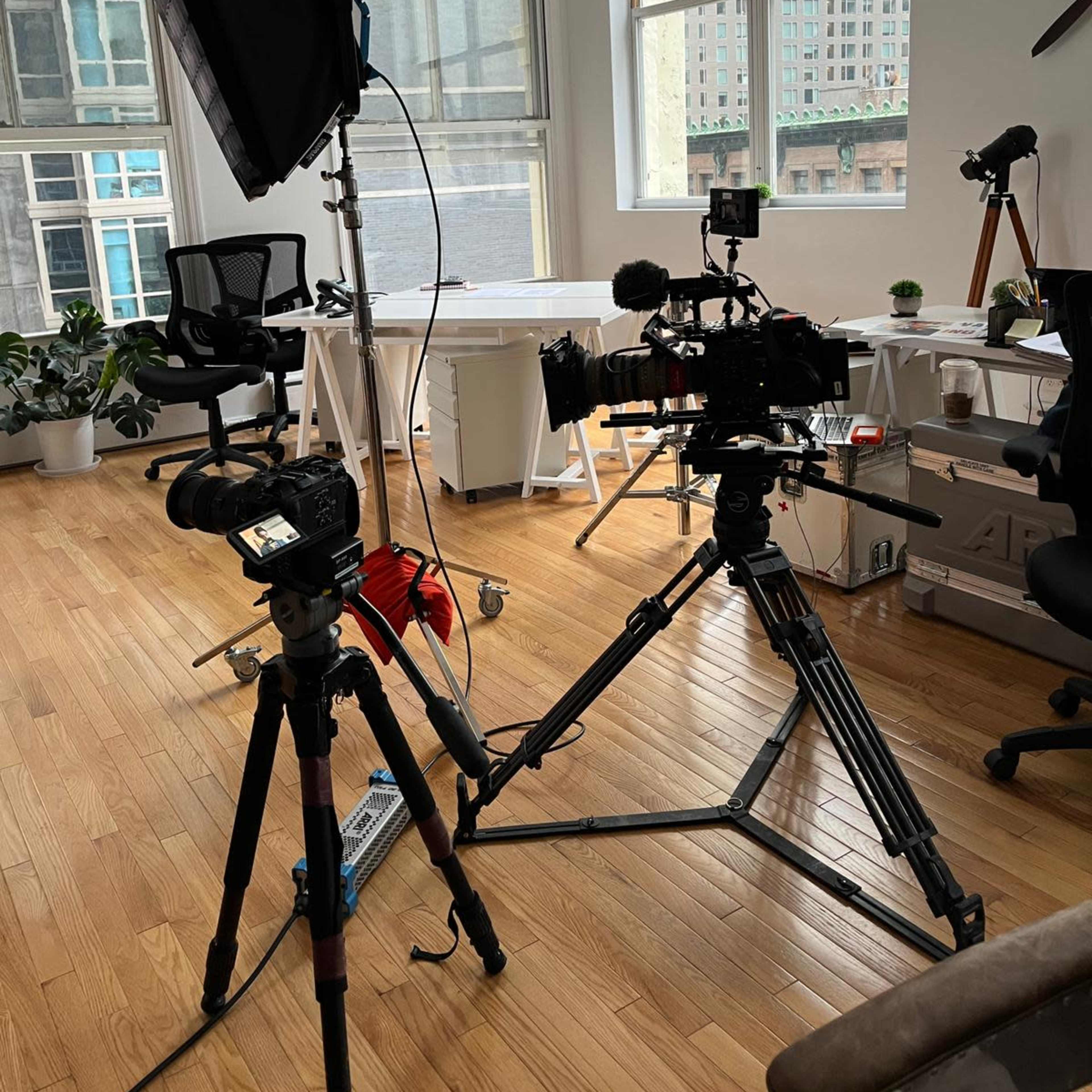 A well-lit office space with video equipment, including tripods, cameras, and softbox lighting, arranged near a large window.