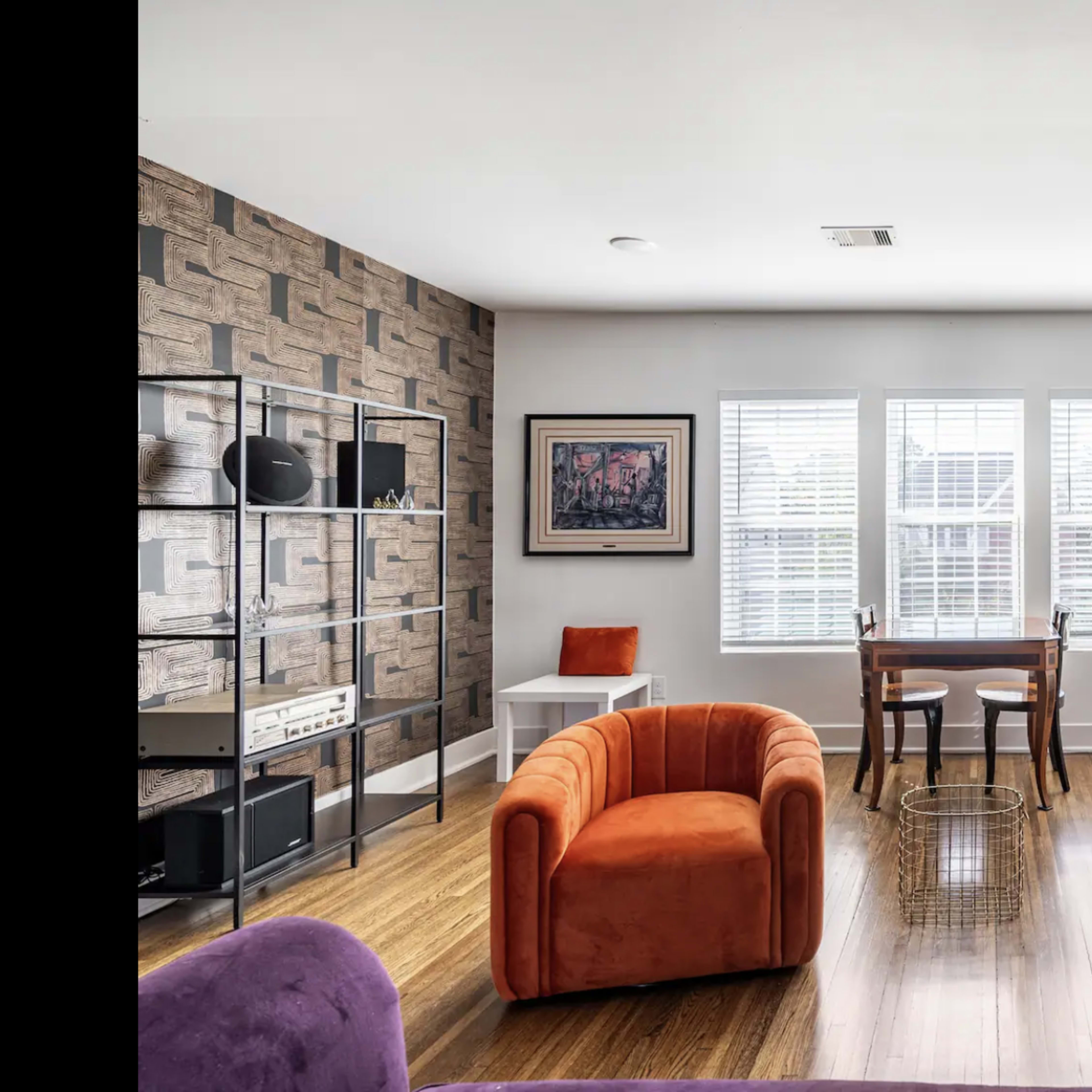 A modern living room with two orange armchairs, a wooden dining table, and large windows allowing natural light to fill the space.