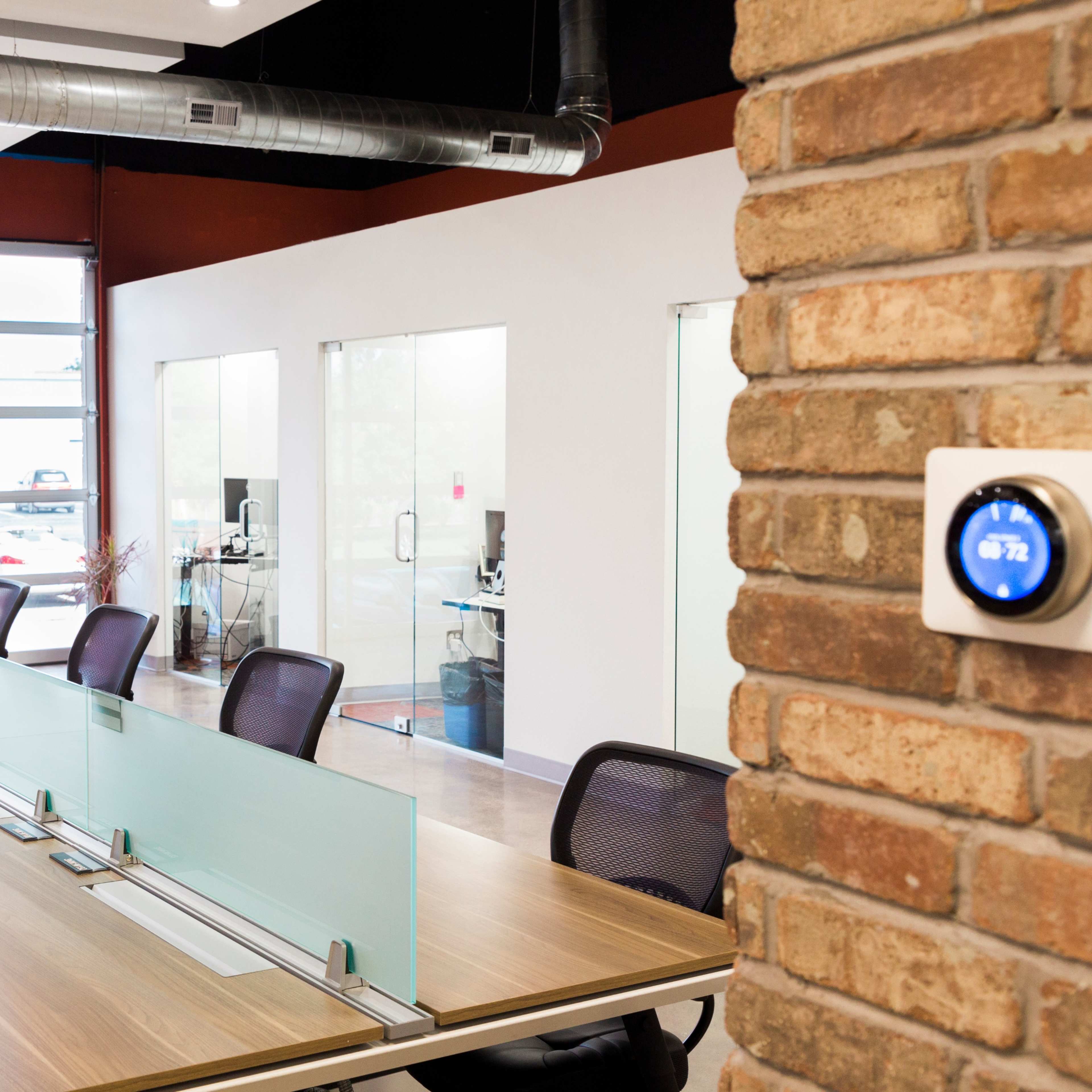 A modern office space features a long table with glass partitions, surrounded by ergonomic chairs, adjacent to large windows and a brick wall with a thermostat.