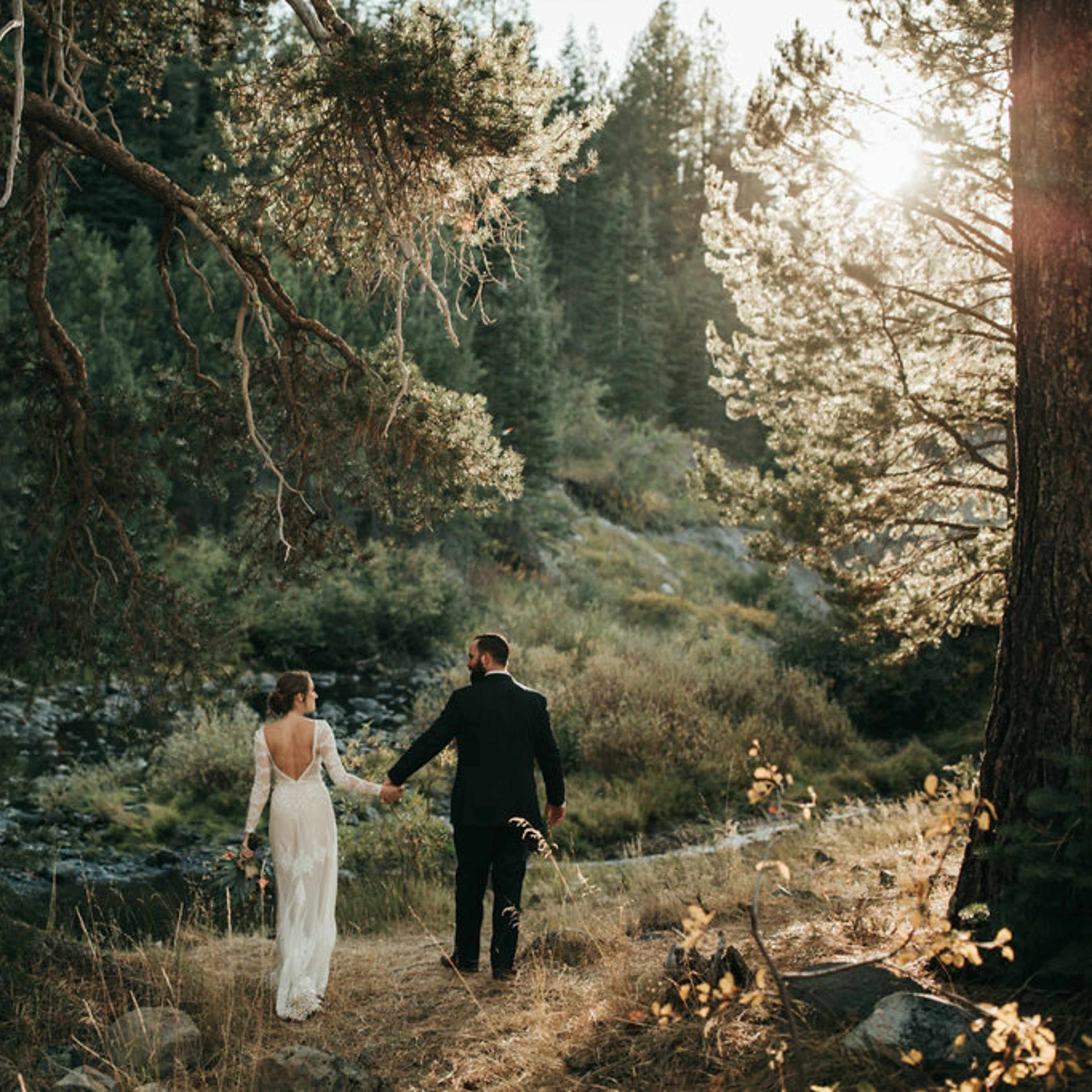 A bride and groom walk hand in hand along a forested path beside a stream, with sunlight filtering through the trees.