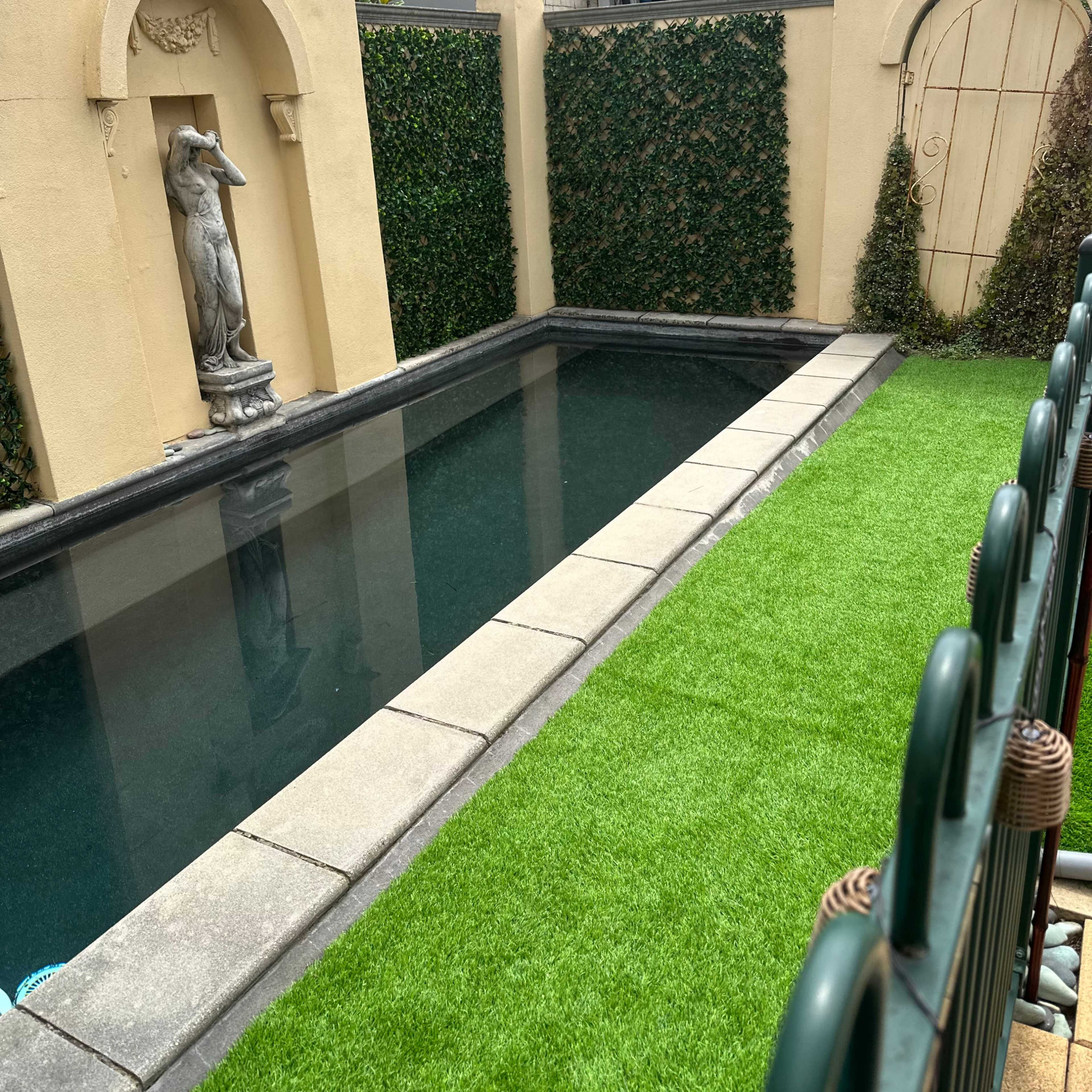 A rectangular swimming pool bordered by stone tiles, with a statue at one end and artificial grass alongside the pool enclosed by a wall.