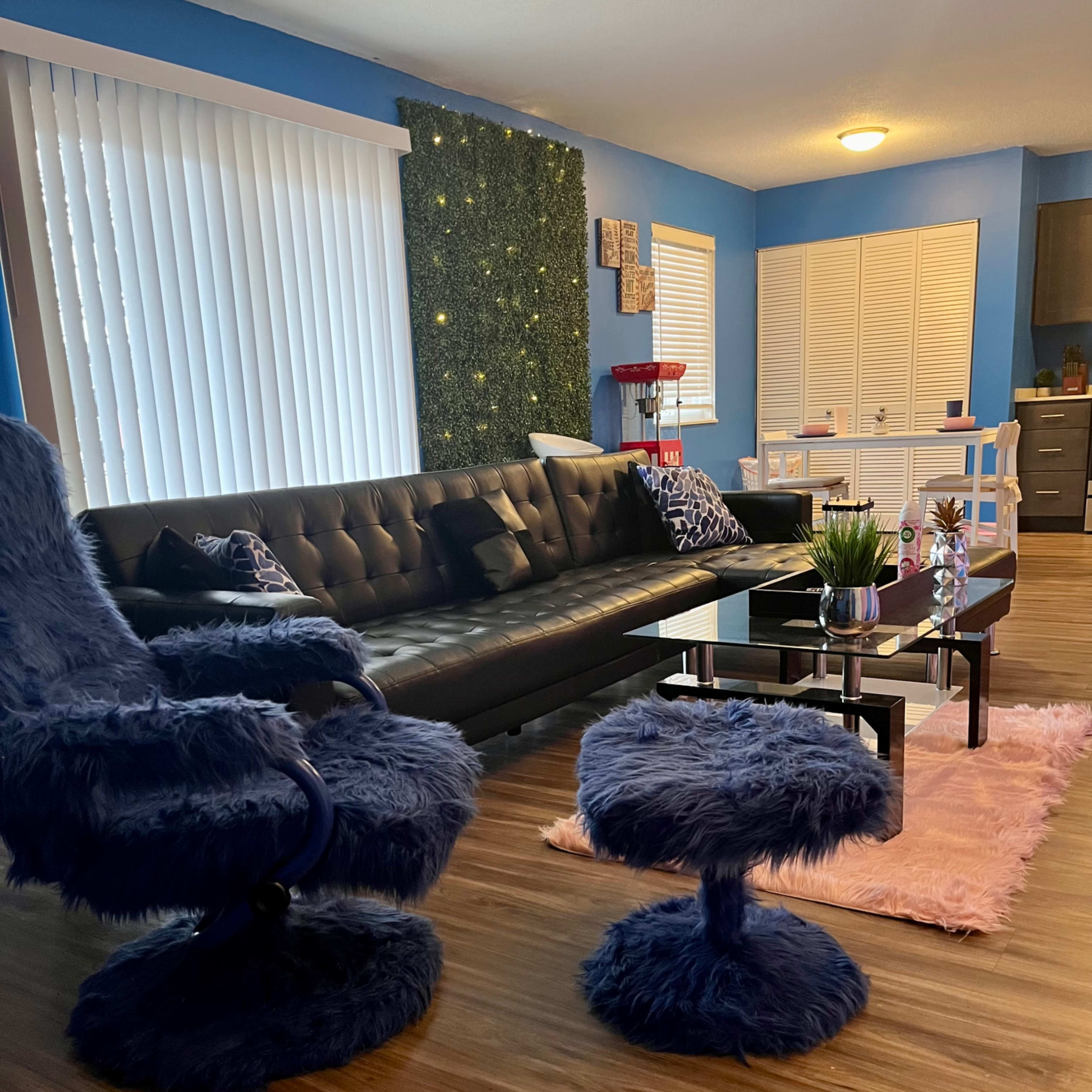 The living room features a black sofa, a blue, fuzzy armchair, and a matching ottoman, with wooden flooring and decorative elements on the walls.