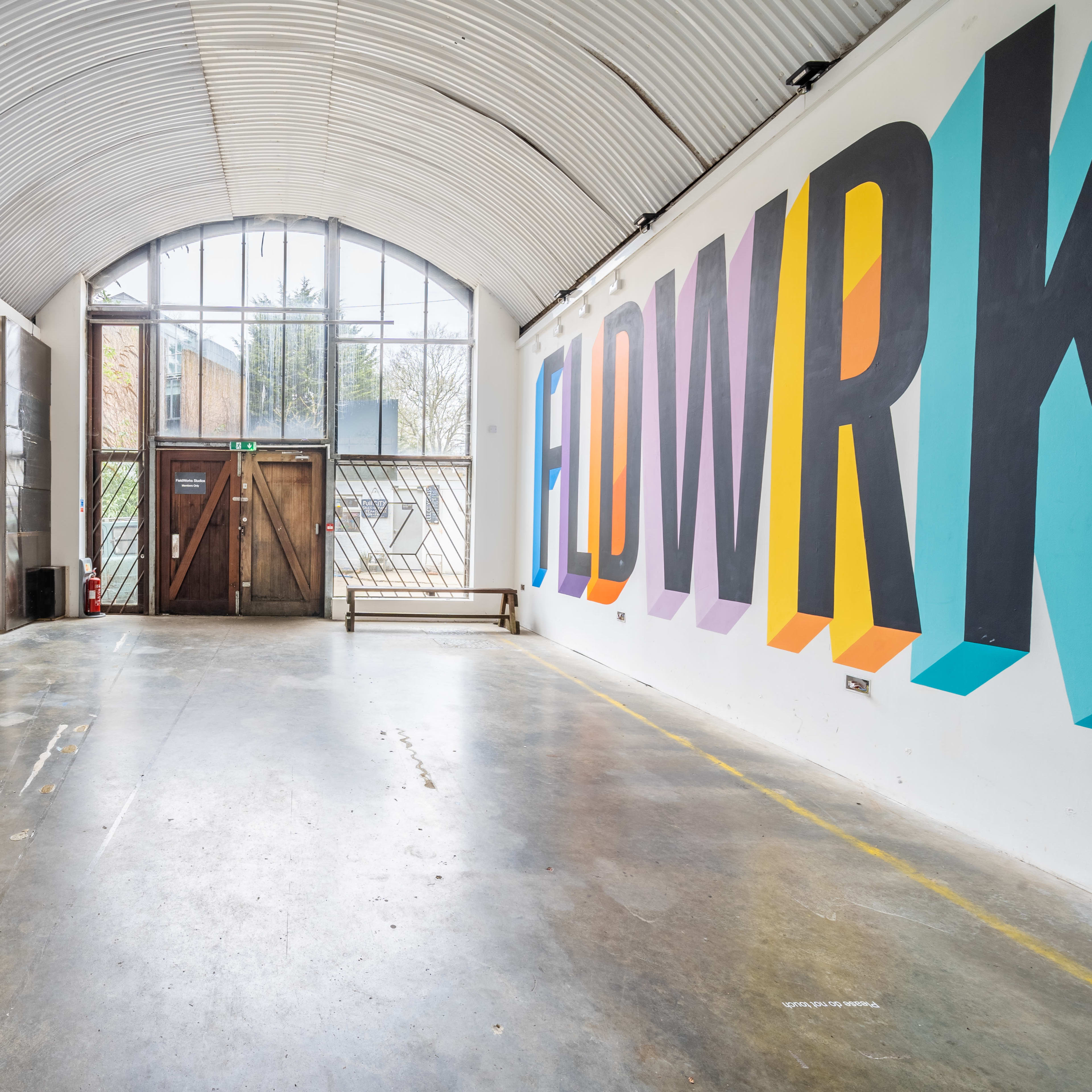A spacious, minimalist interior with a large colorful mural that reads "FIDWRK" on one wall and a prominent glass door at the entrance.