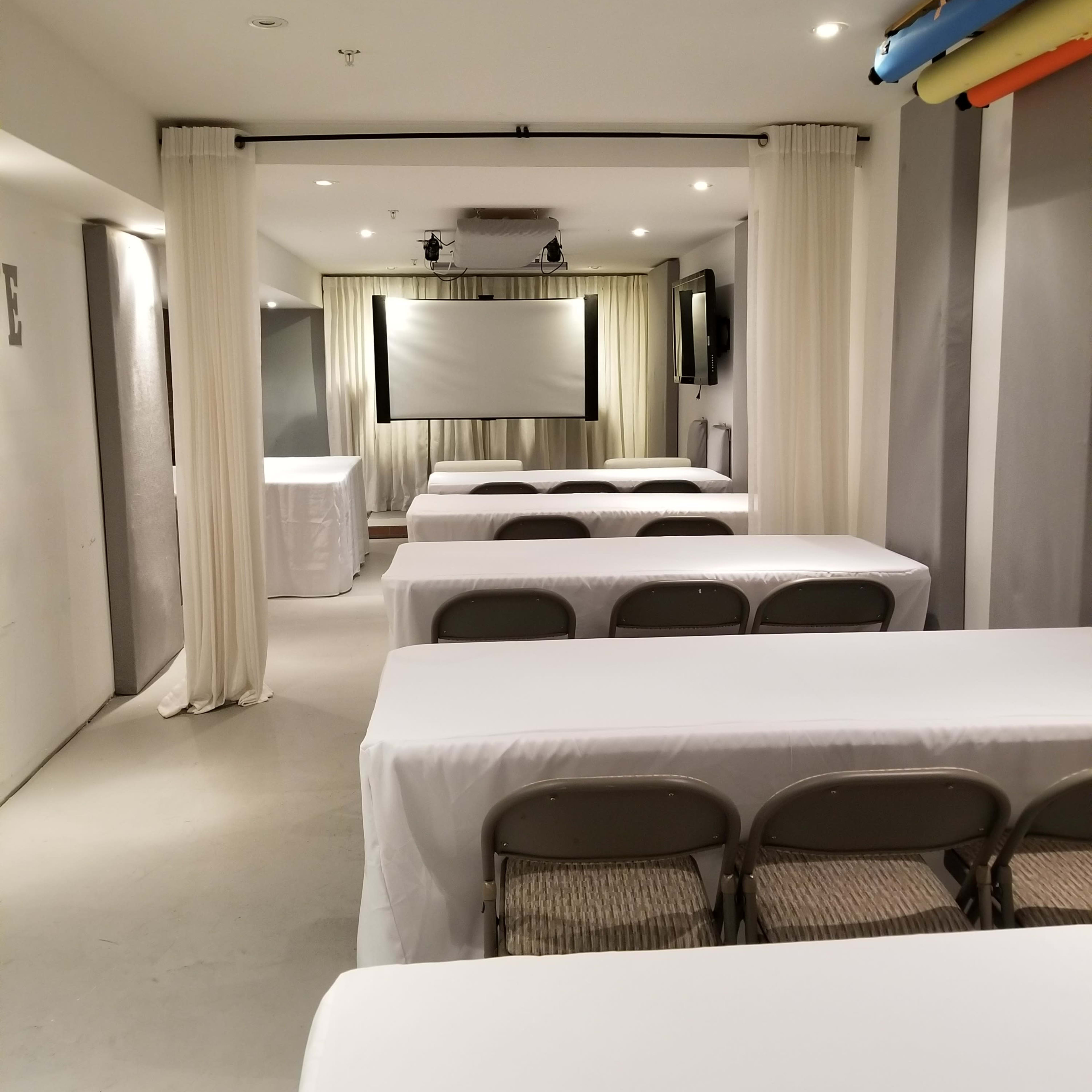 A conference room features several rows of tables covered with white tablecloths, facing a projector screen at the front.
