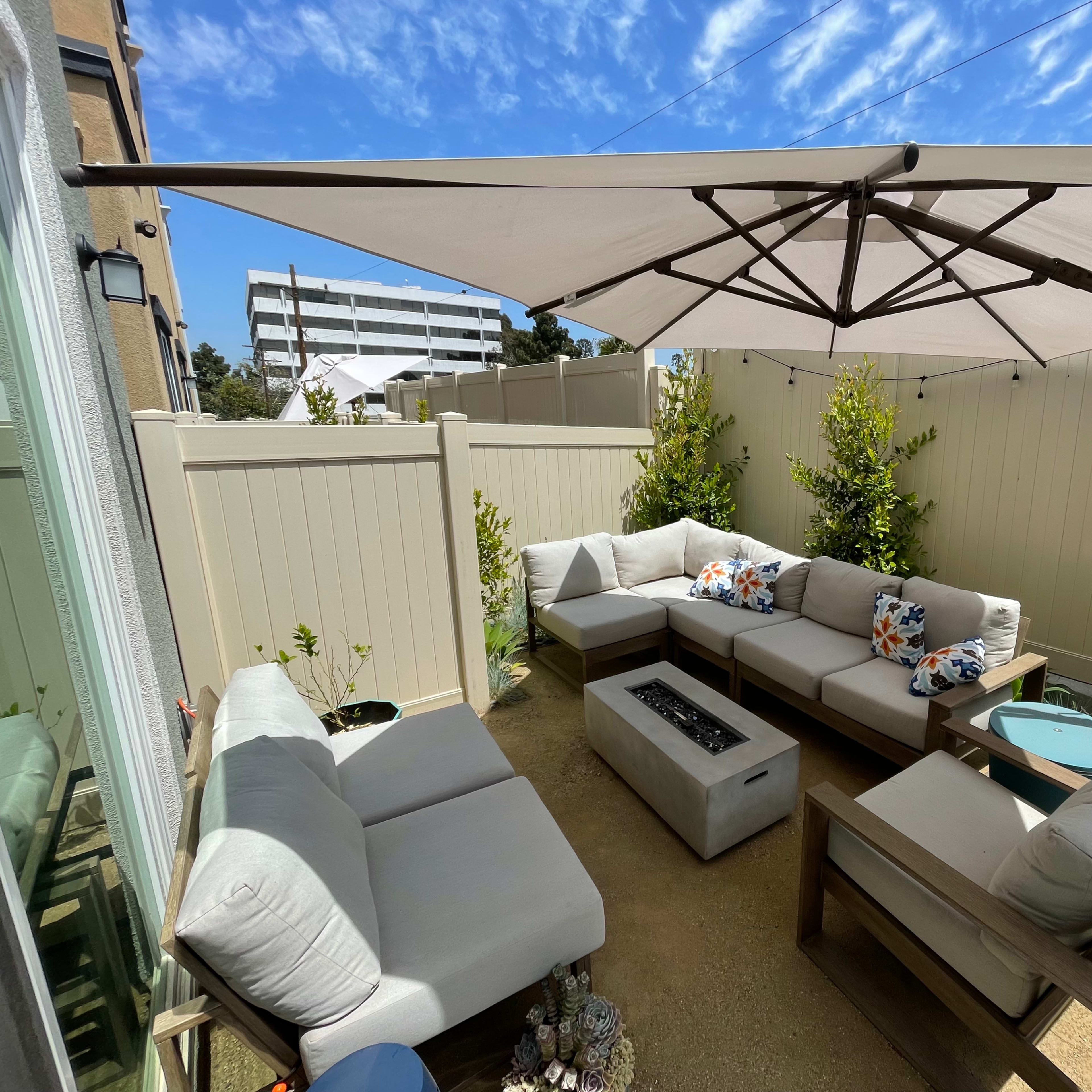 A patio area features a sectional sofa under a large umbrella, with a fire pit in the center and surrounding greenery.
